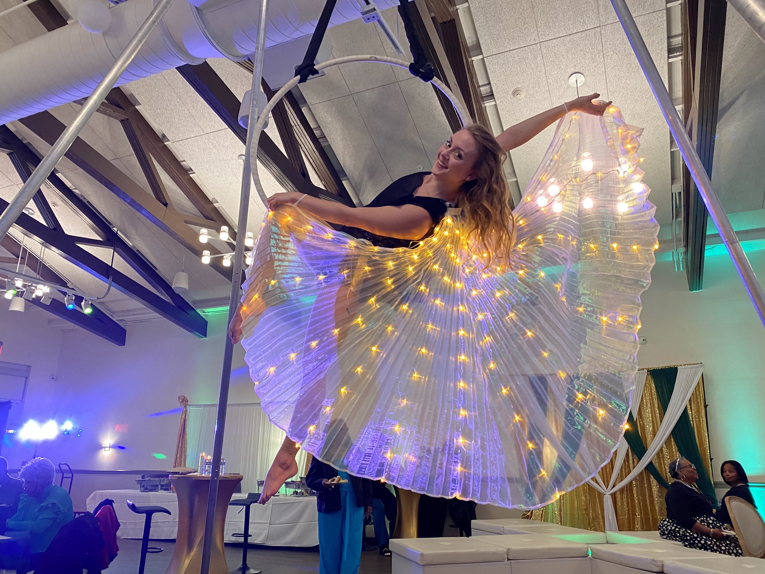 A woman performing an aerial dance on a hoop decorated with LED lights, in a colorful indoor venue with mood lighting and decorations.