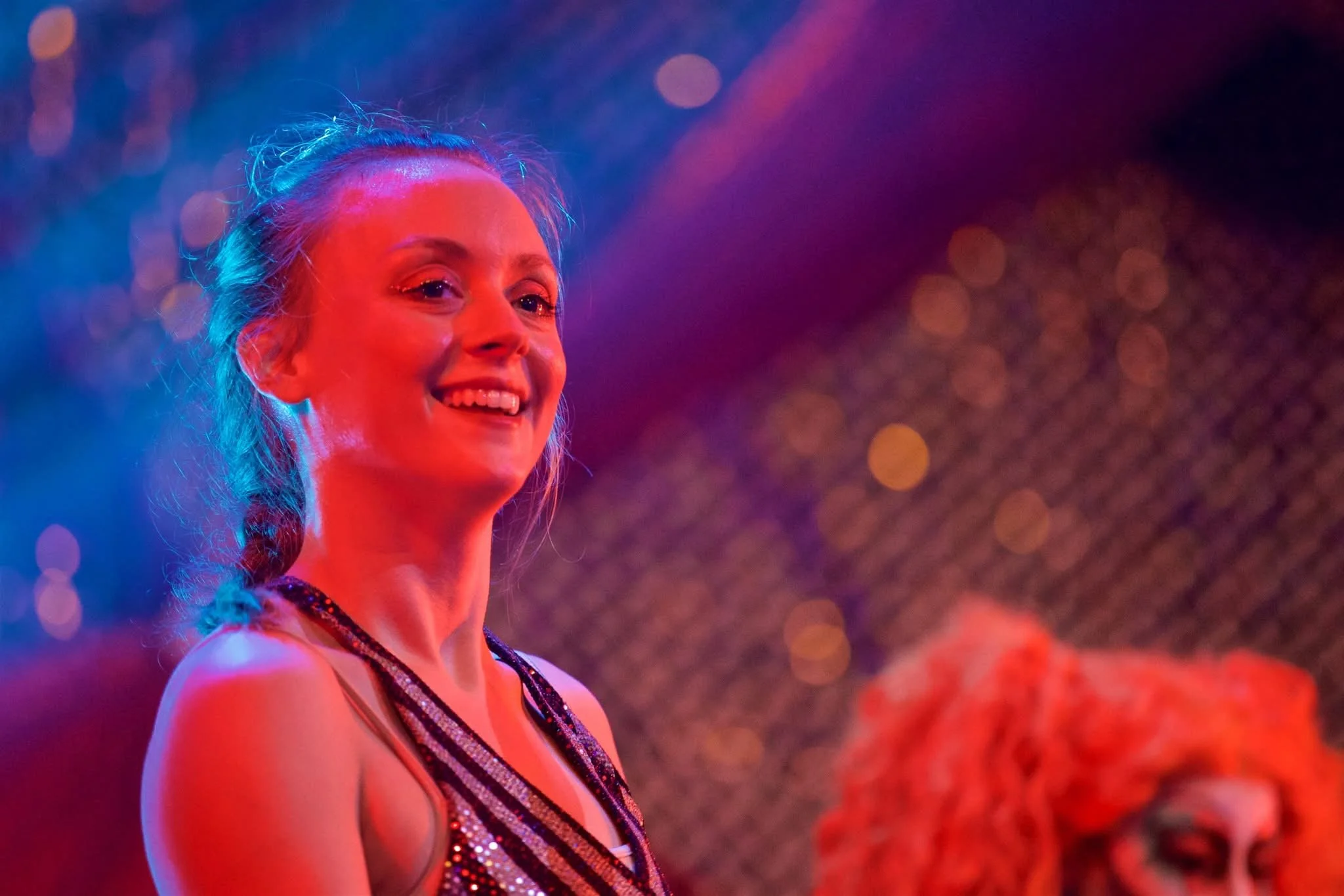 A woman smiling, with colorful stage lighting and bokeh background, wearing a sparkly dress.