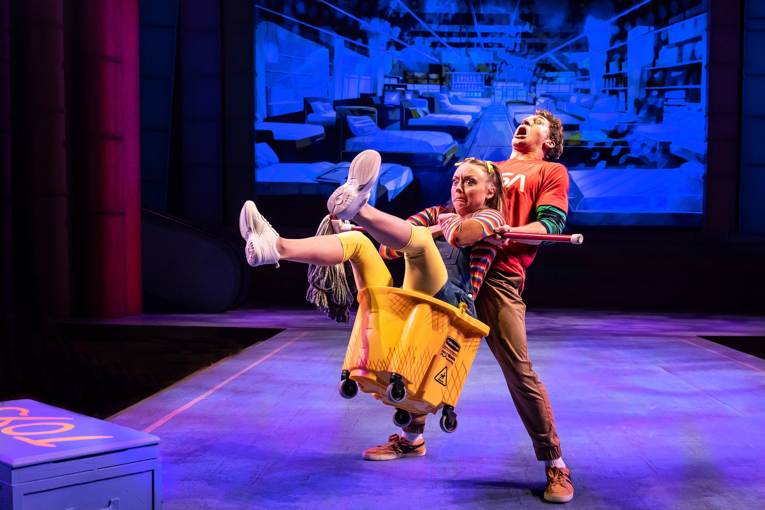 A theatrical stage performance featuring two actors, one male and one female, in a comedic scene. The female actor is sitting in a yellow shopping cart and appears surprised or distressed, while the male actor stands behind her, holding a broom and m
