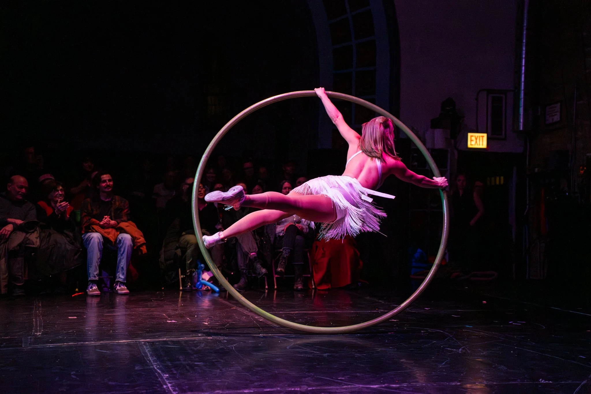 A performer in a white fringed costume performing on a Cyr wheel on stage, with an audience watching in the dark background.