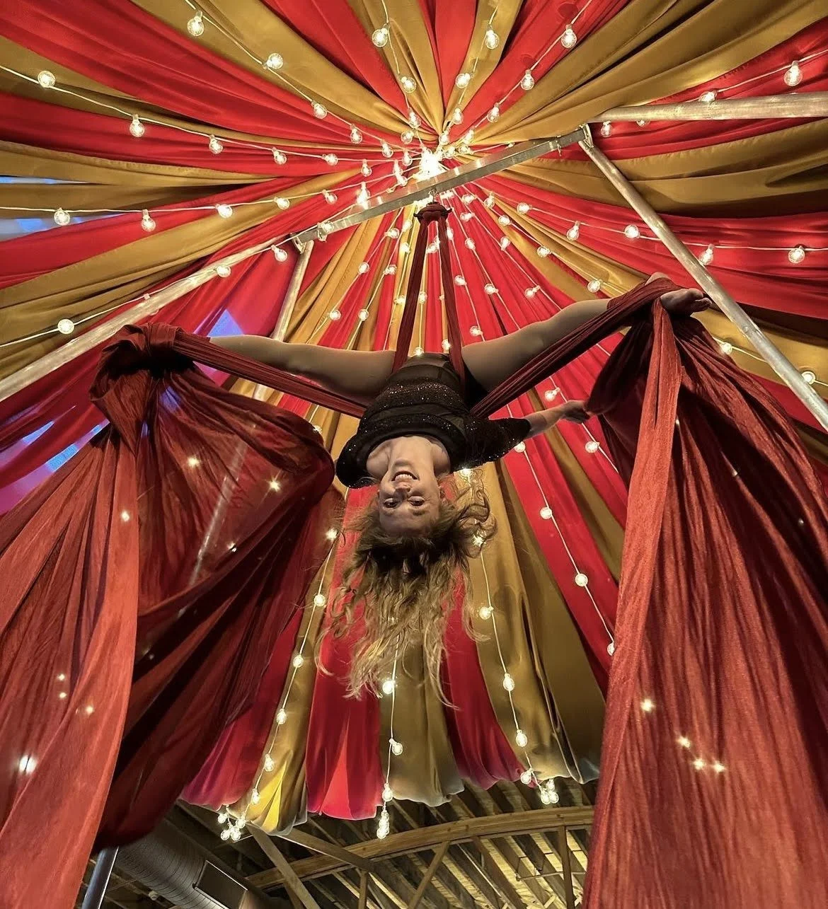 A performer hanging upside down on red and gold draped fabric with string lights overhead, smiling during a circus or stage act.