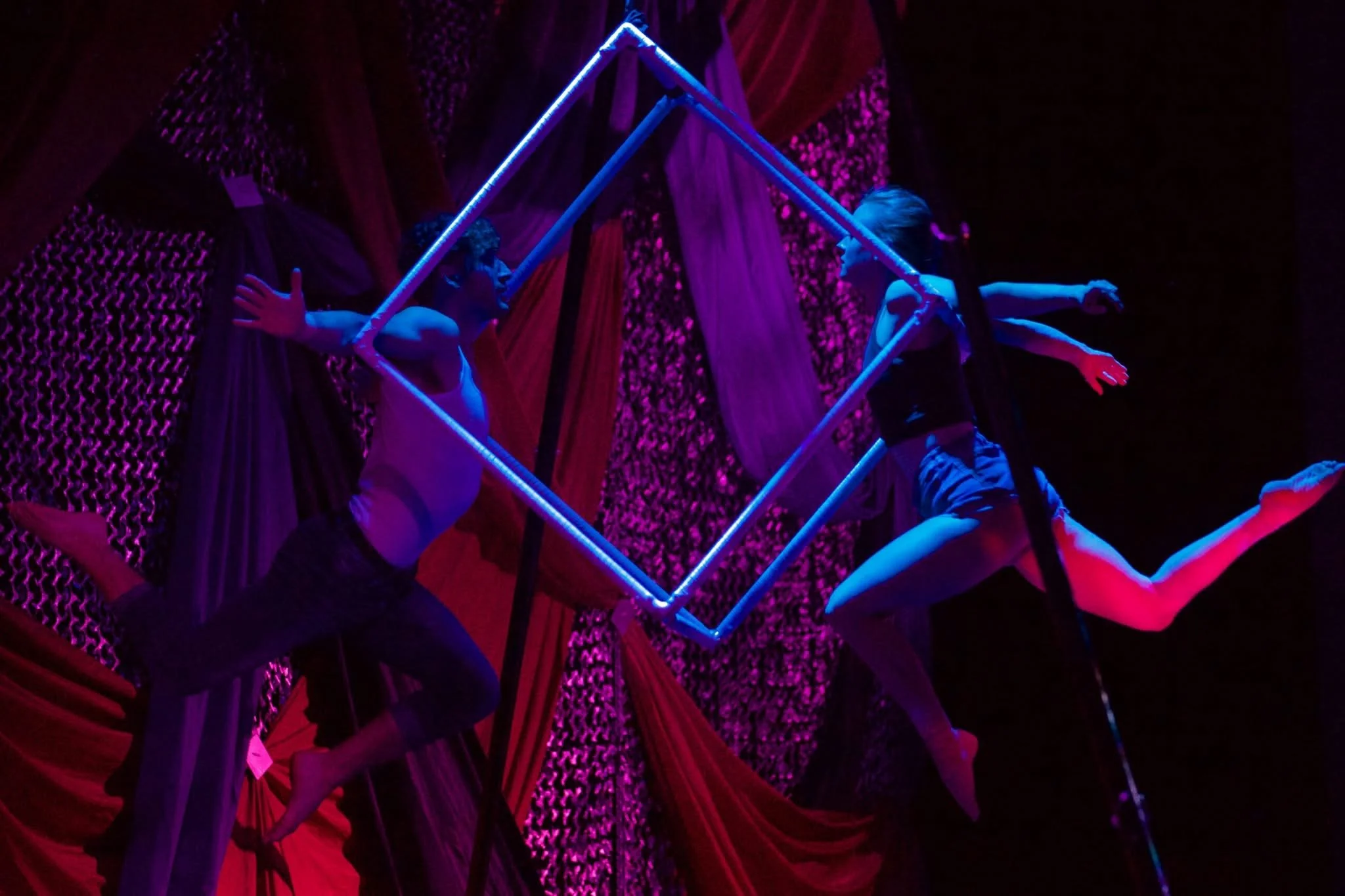 Two circus performers in black shorts and leotards perform an aerial act with long poles inside a tent, illuminated by purple and pink stage lights.