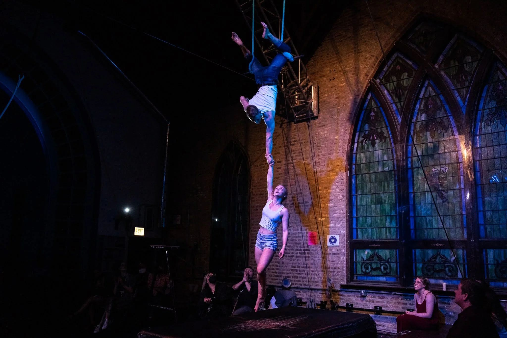 Two acrobats performing an aerial act inside a brick-walled venue with stained glass windows. The female acrobat is on the ground, reaching up with one hand as the male acrobat hangs upside-down from a hanging apparatus above her, gripping her hand.