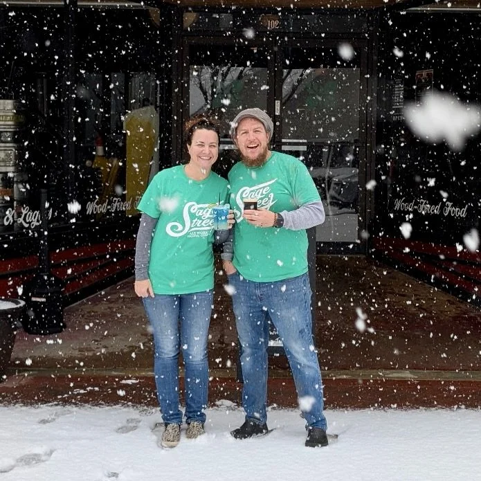 Two smiling people in teal shirts standing outside in falling snow in front of a restaurant with snow on the ground.
