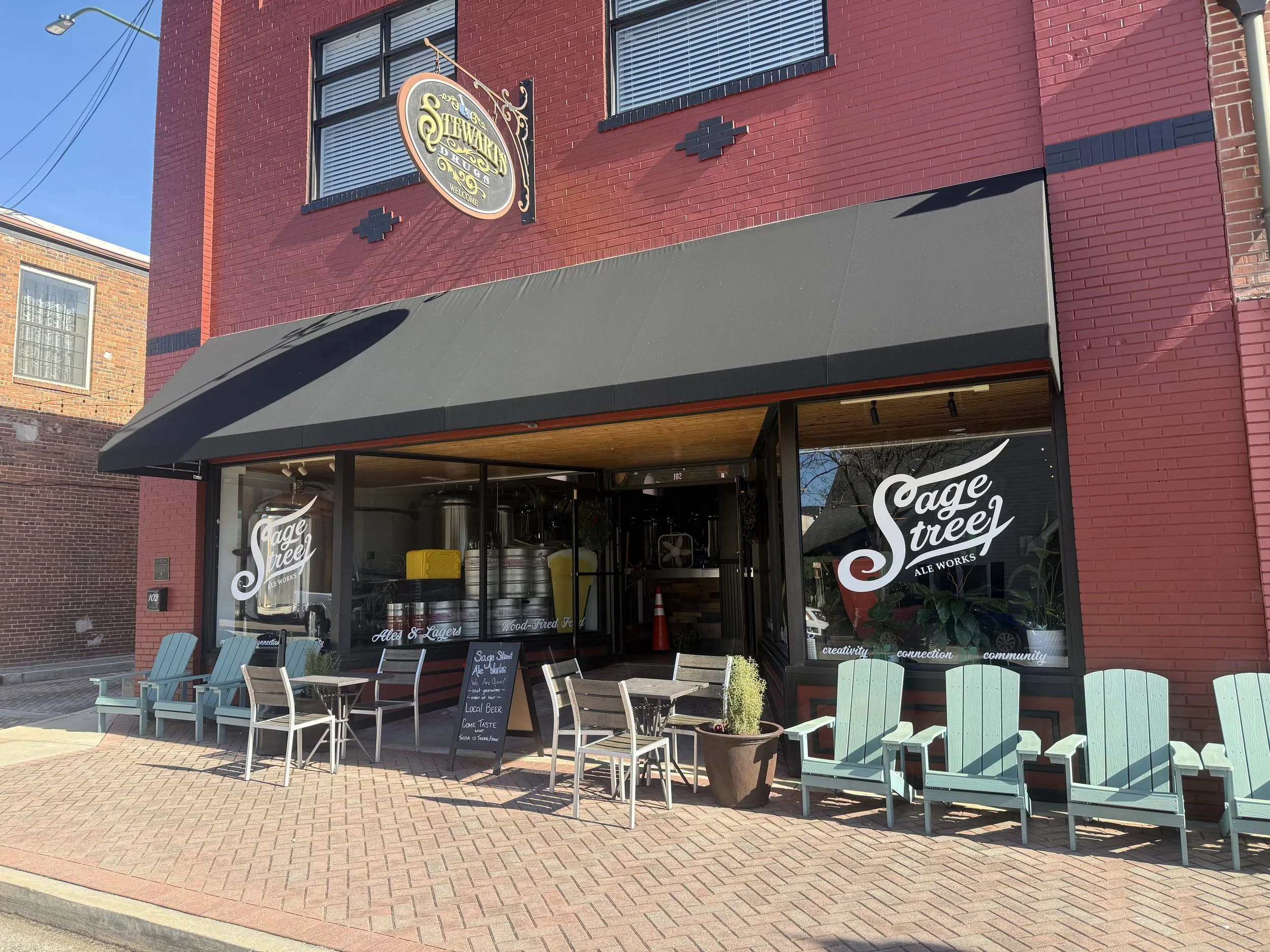 Front view of Sage Street Ale Works brewery with outdoor seating, large windows, a black awning, and brewery tanks visible inside