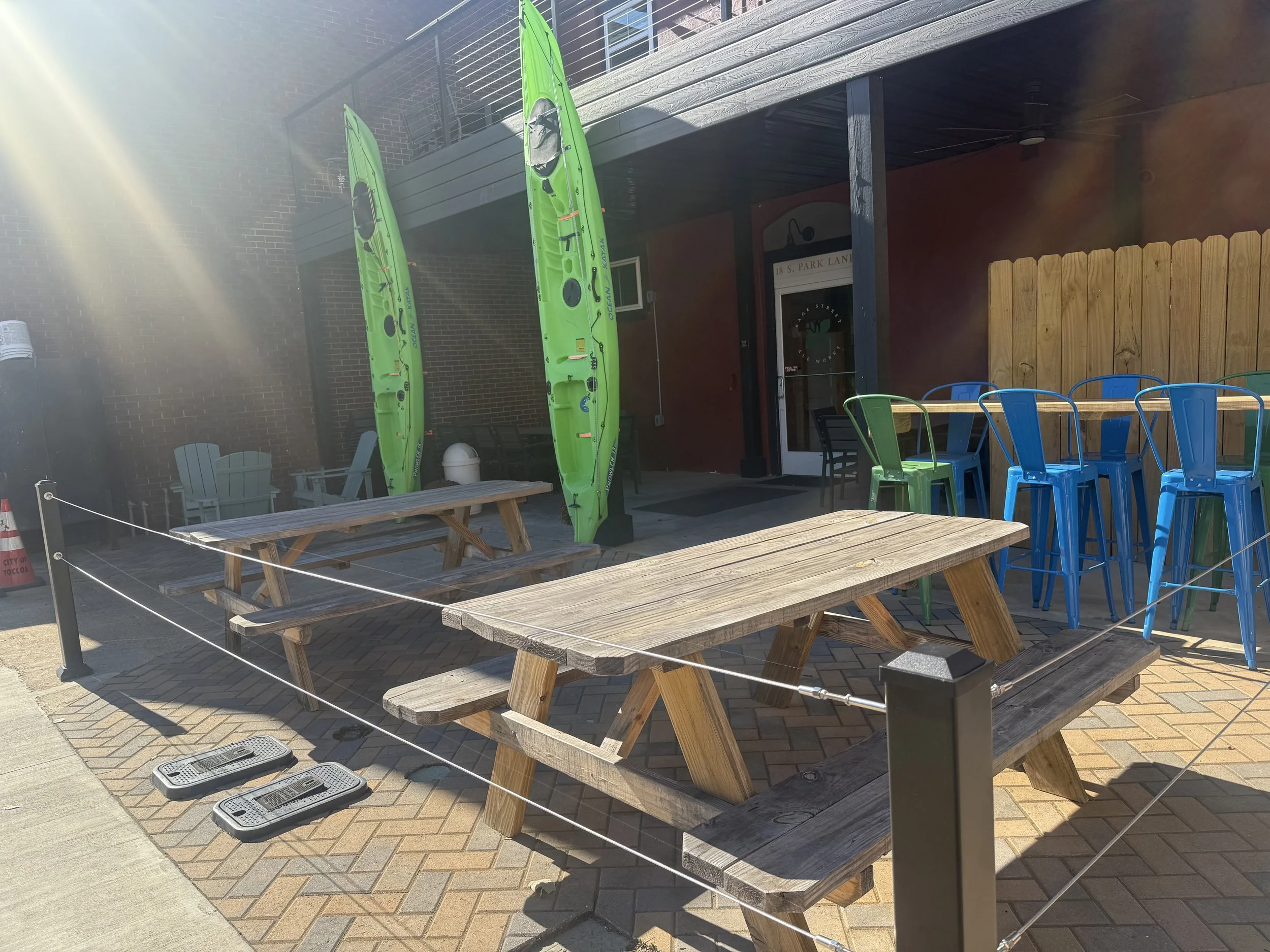 Outdoor seating area with wooden picnic tables, plastic chairs in blue and green, and two green kayaks upright against a brick wall. Sunlight streaming over the scene and a wooden fence in the background.