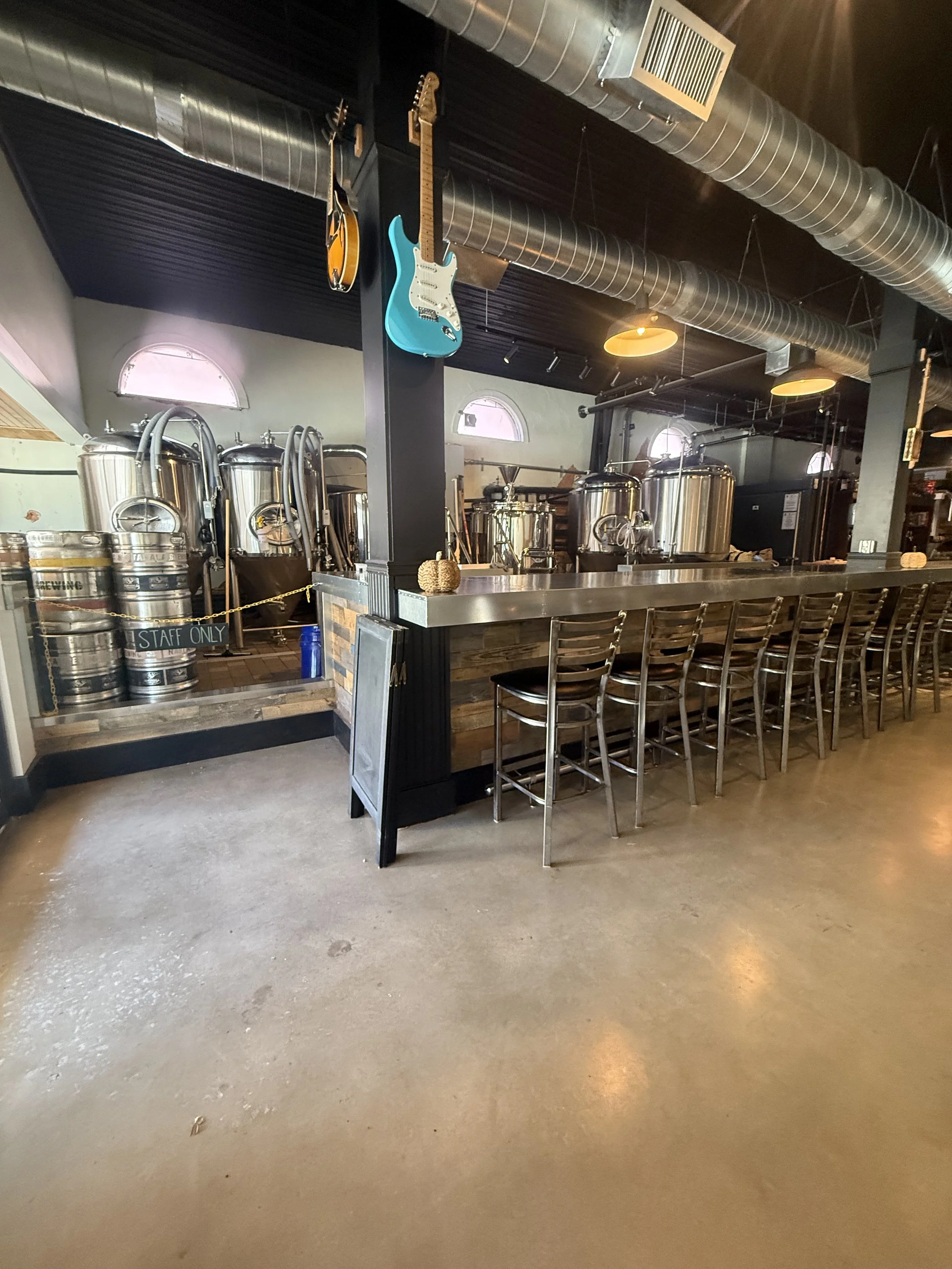 Bar area with high chairs, beer kegs, brewing equipment, and decorative guitars hanging from the ceiling in a brewery or pub.