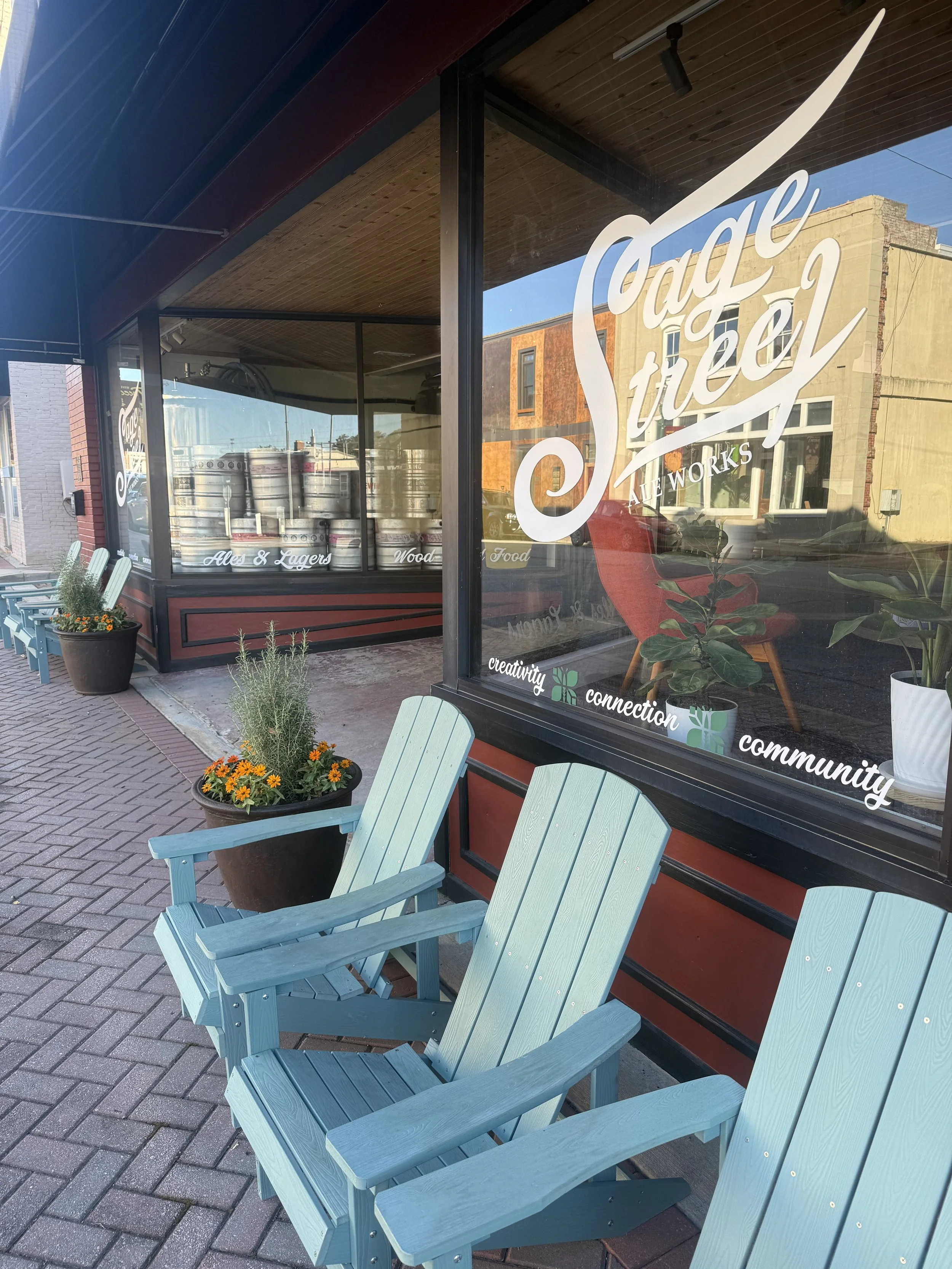 Sidewalk cafe outside a craft beer and food shop with light blue Adirondack chairs, potted plants, and a large window with signage and beer kegs inside.
