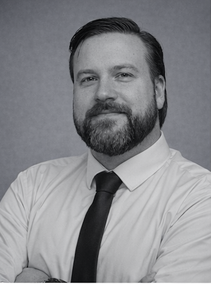 A black-and-white portrait of a man with a beard, wearing a white shirt and dark tie, smiling slightly and looking at the camera.