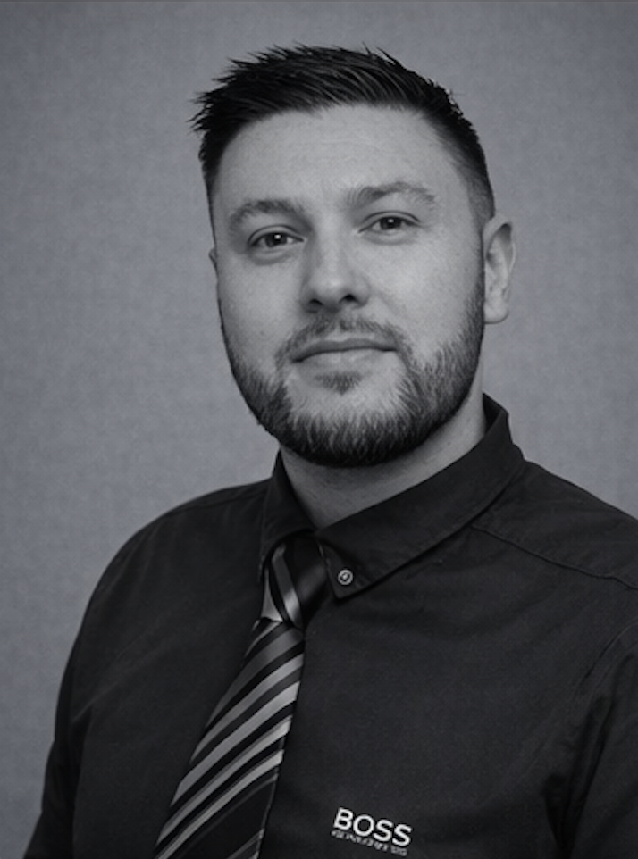 Black and white portrait of a young man with short dark hair, a beard, wearing a black dress shirt with the BOSS logo and a striped tie against a plain background.