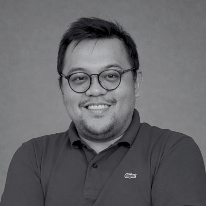 Black and white portrait of a man wearing glasses and a collared shirt, smiling with arms crossed against a plain background.