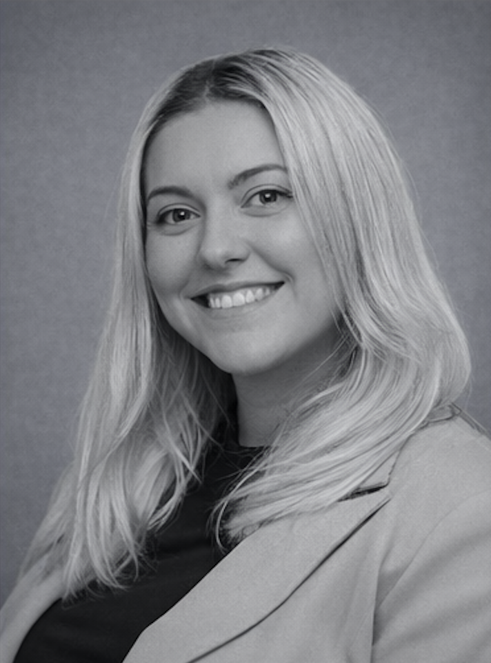 Black-and-white portrait of a smiling young woman with long, wavy blonde hair, wearing a blazer and a dark top, against a plain background.