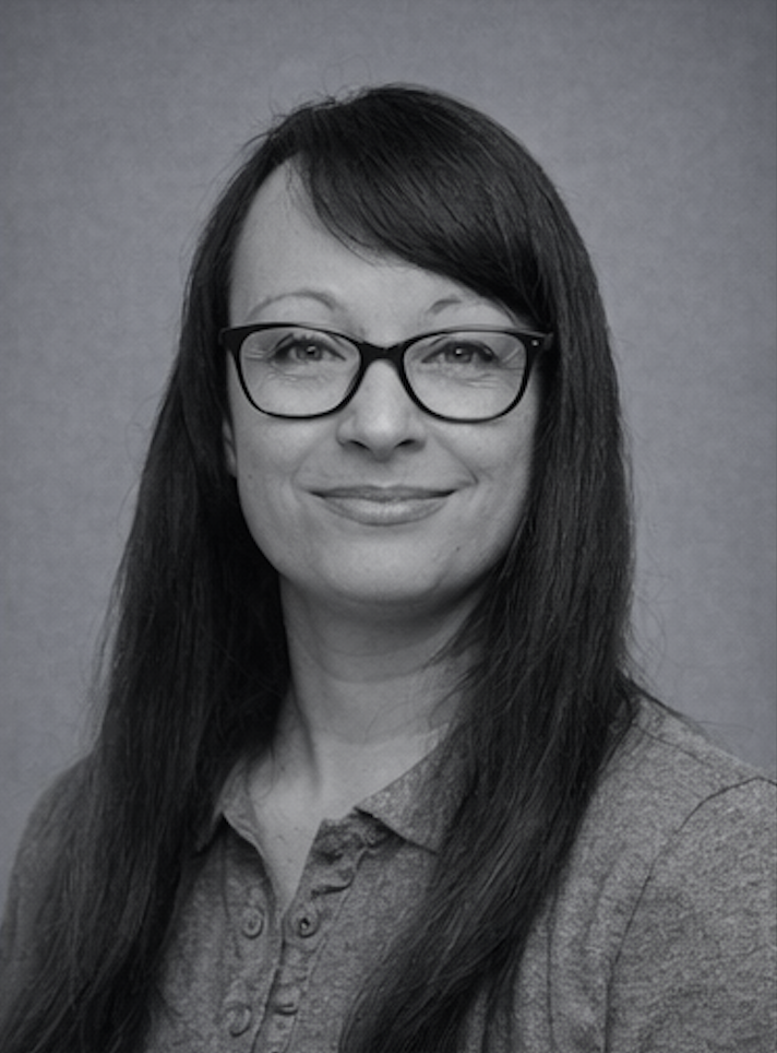 A woman with long dark hair, wearing glasses and a button-up shirt, smiling at the camera in a professional headshot.