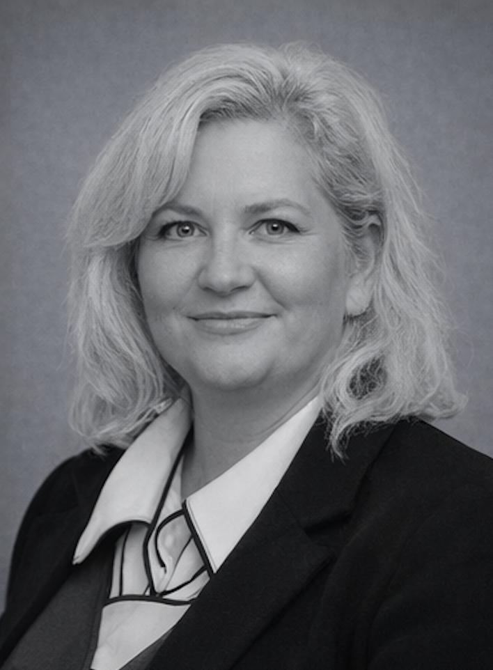 Black and white professional portrait of a middle-aged woman with shoulder-length wavy blonde hair, wearing a dark blazer and a white collared shirt, against a plain background.