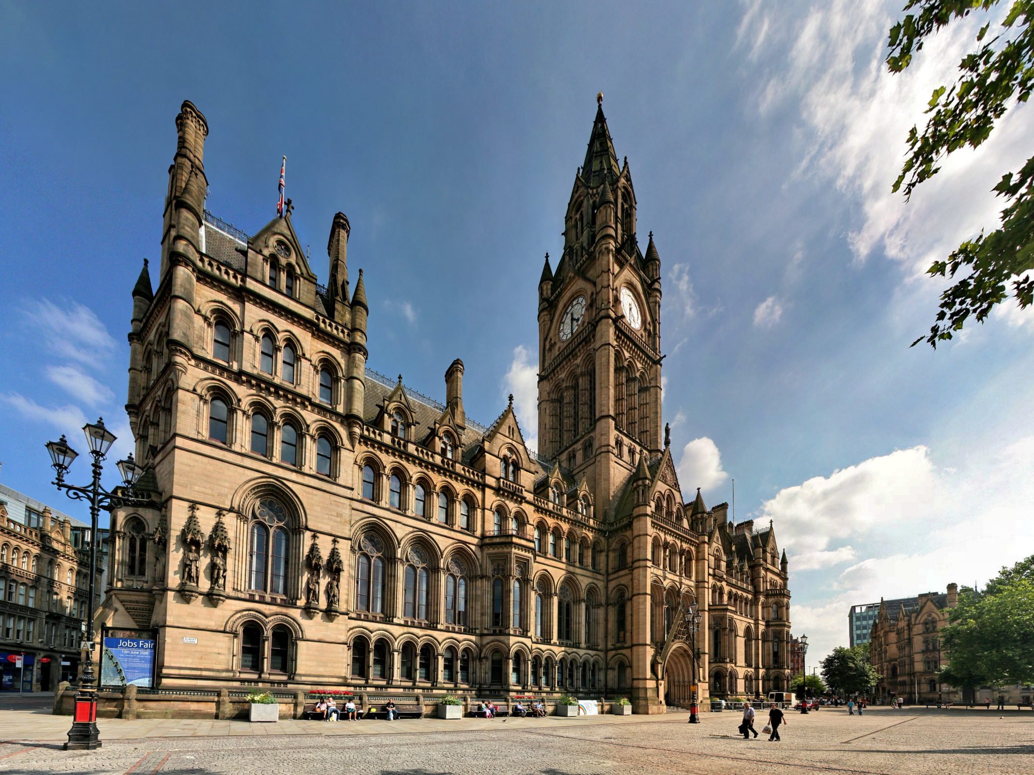 A historic Gothic-style building with a tall clock tower under a partly cloudy sky, located in a city square with a few people walking and sitting.