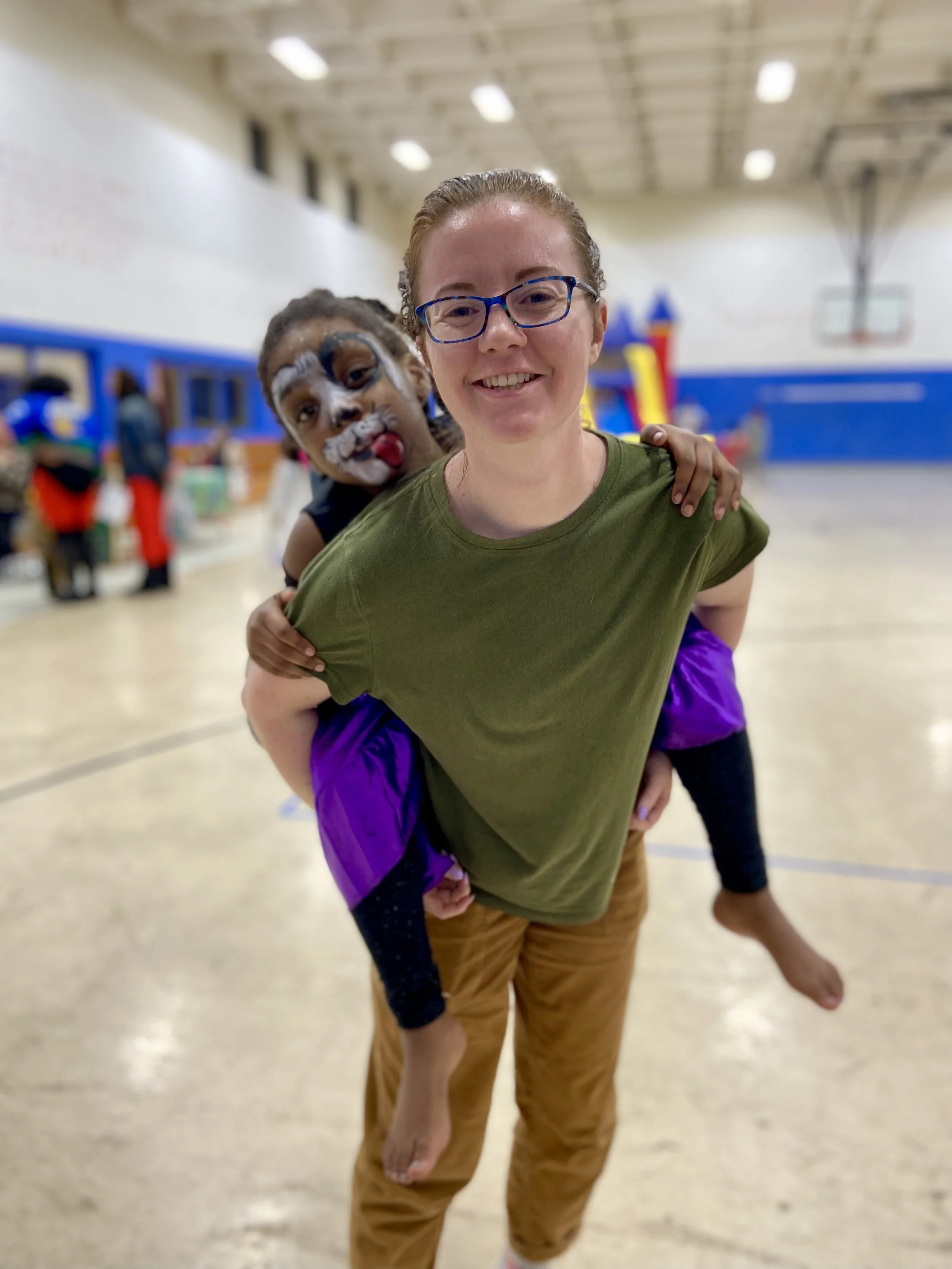 A smiling woman with glasses carrying a young girl on her back in a gymnasium, with some children and play equipment in the background.