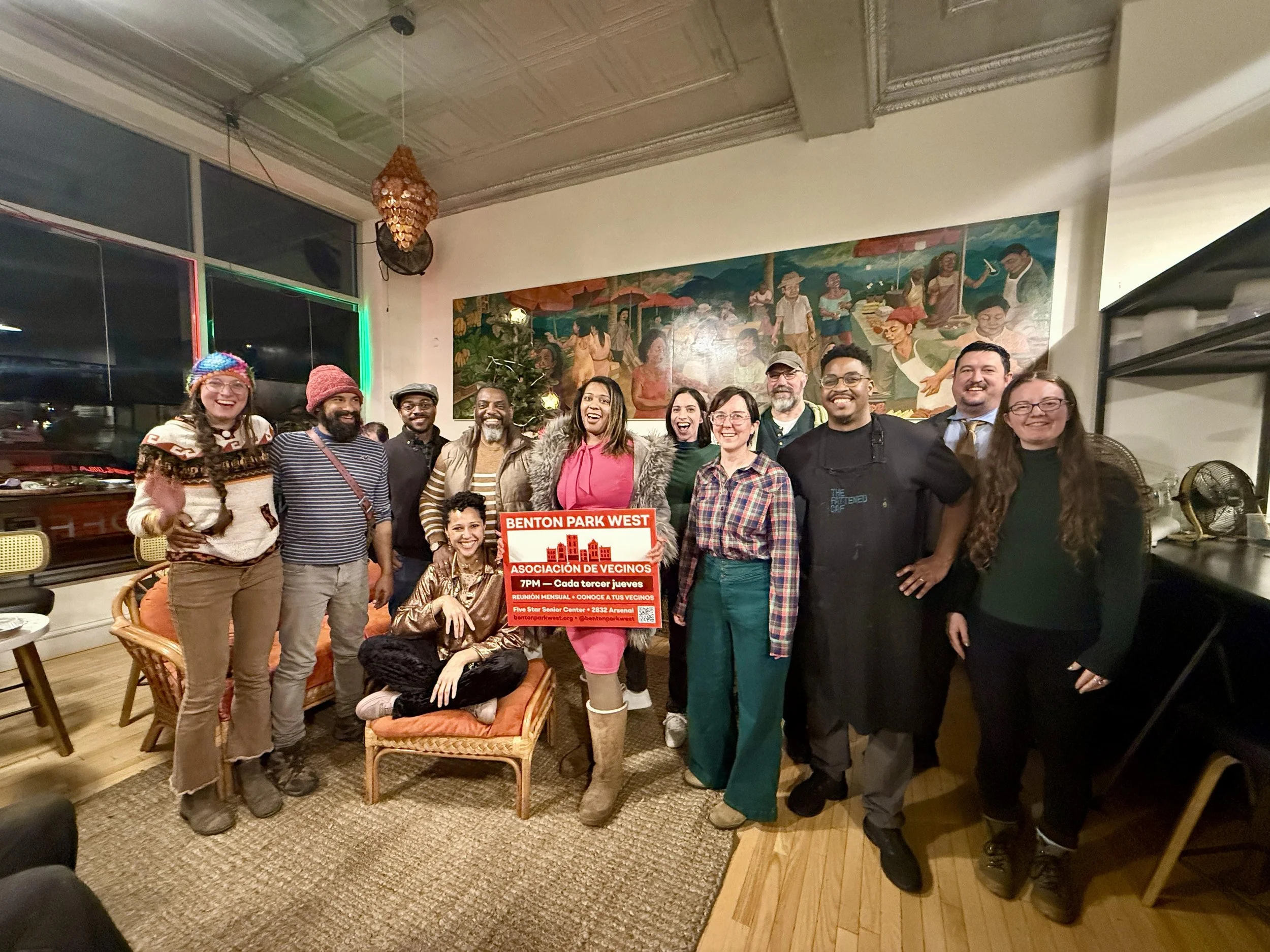 A diverse group of people stands together smiling for a photo in a warmly lit room. They are holding a red and white sign that reads "Benton Park West Asociación de Vecinos 7PM - Cada tercer jueves." The background features a large colorful mural and a window with multicolored lights.