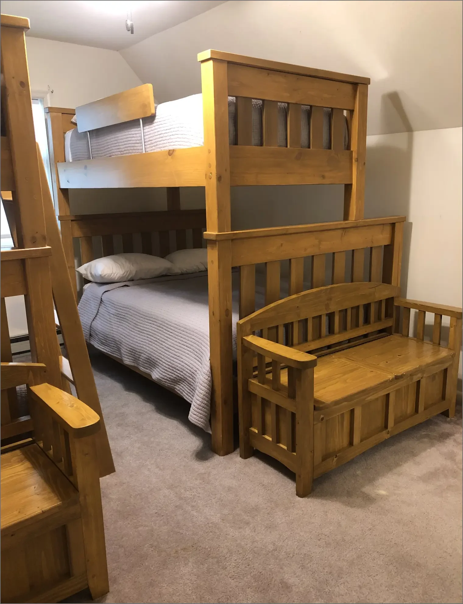 A room with a wooden bunk bed with a white mattress and pillows, a wooden bench at the foot of the bed, and part of a wooden chair visible in the foreground.