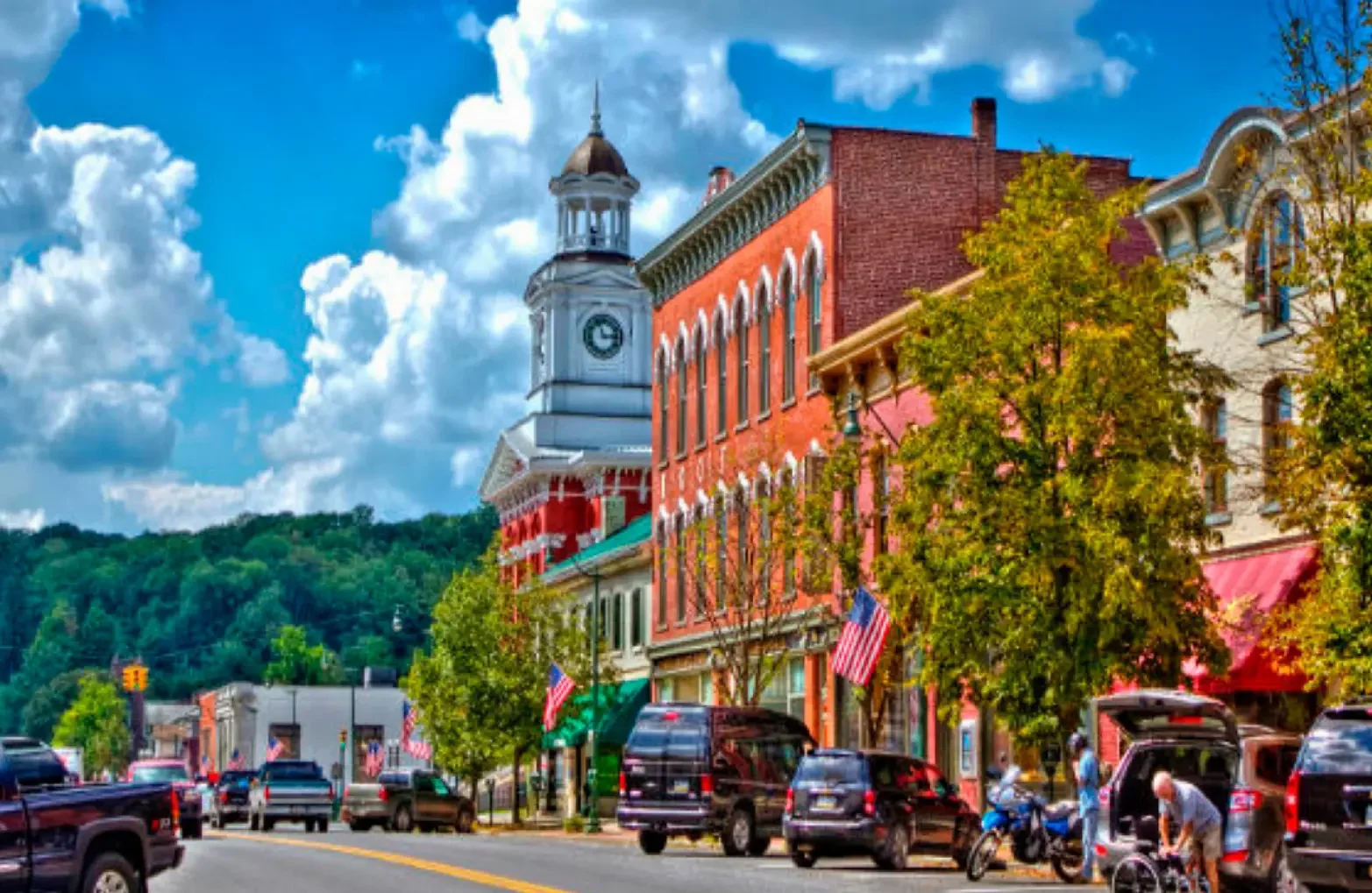 Main street in a small town with colorful buildings, American flags, trees, parked cars, and a mountain with a blue sky and clouds in the background.