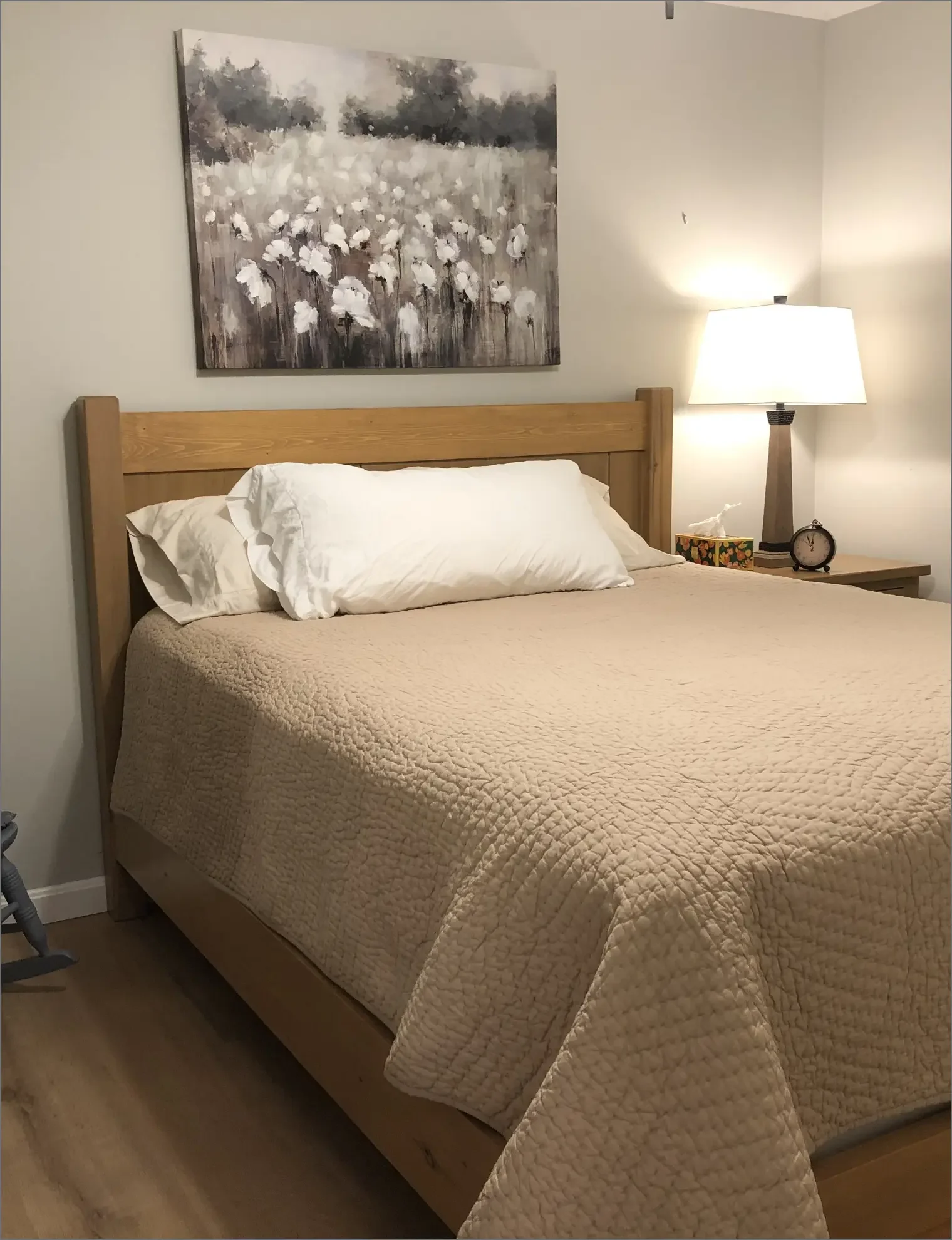A neatly made bed with a wooden headboard, white pillows, and a beige quilt, situated beside a wooden nightstand with a lamp, a clock, and a tissue box. A painting of white flowers on a gray background hangs above the bed.