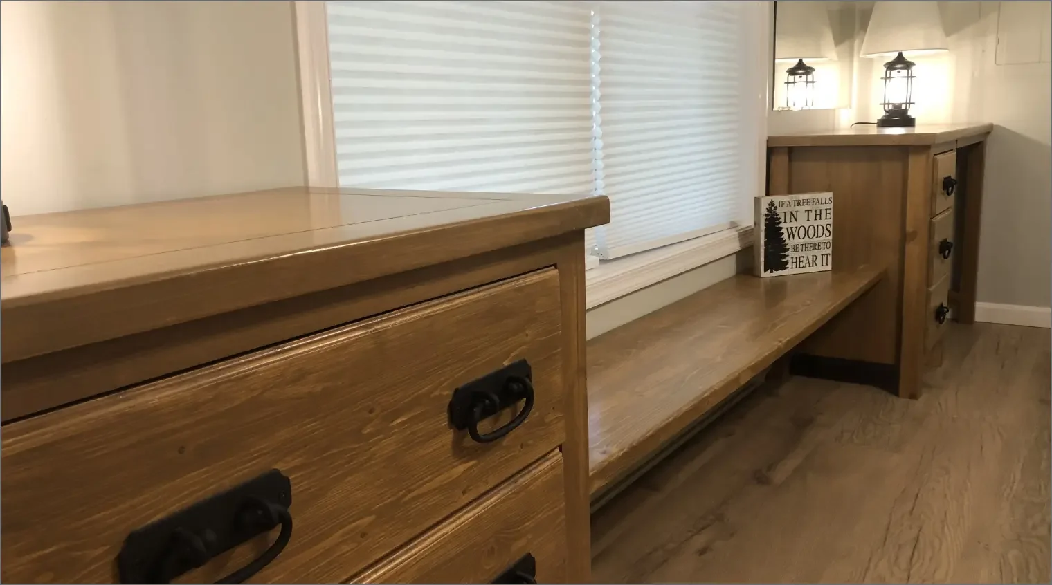 Wooden furniture including a dresser and a long bench with a small sign reading 'If a tree falls in the woods, be there to hear it' under two lantern-style lamps in a room with hardwood floors and horizontally slatted window blinds.