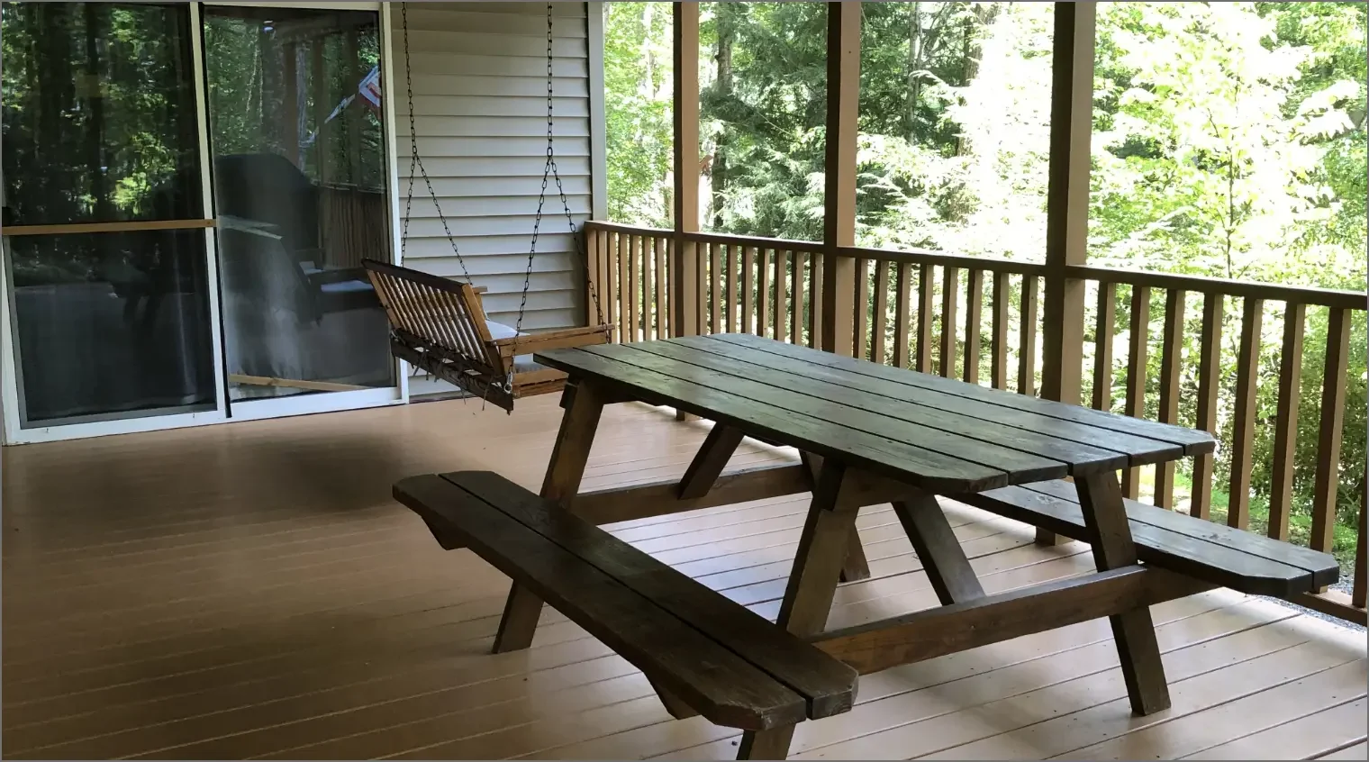 Wooden porch with a picnic table and a porch swing, overlooking a lush green wooded area.