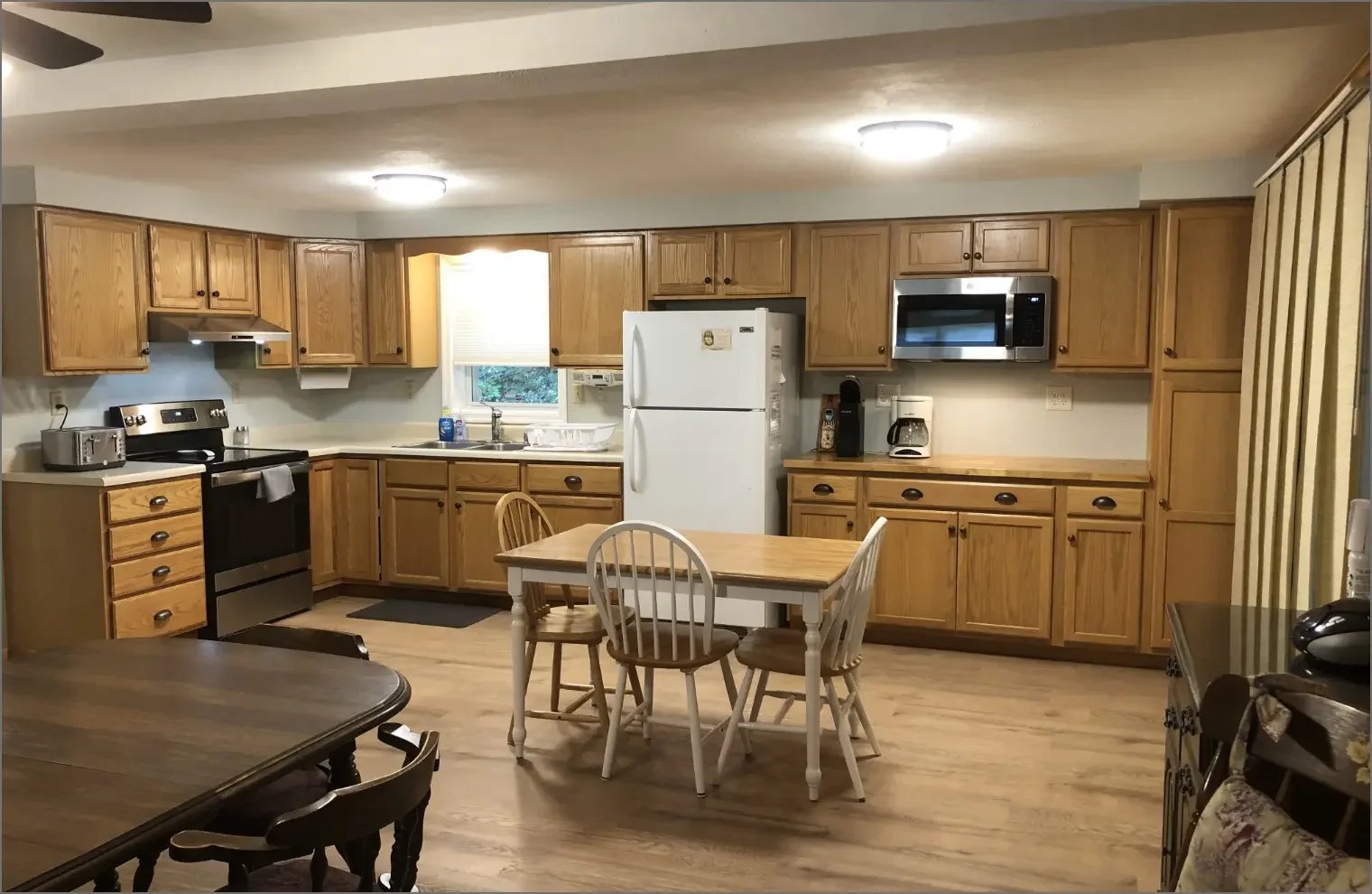 Kitchen with wooden cabinets, white refrigerator, microwave, coffee maker, toaster, sink under window, dining table with four chairs, and hardwood floor.
