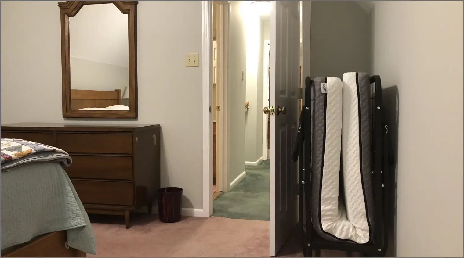 Part of a bedroom with a bed, dresser, mirror, and a folding bed in a carry bag.