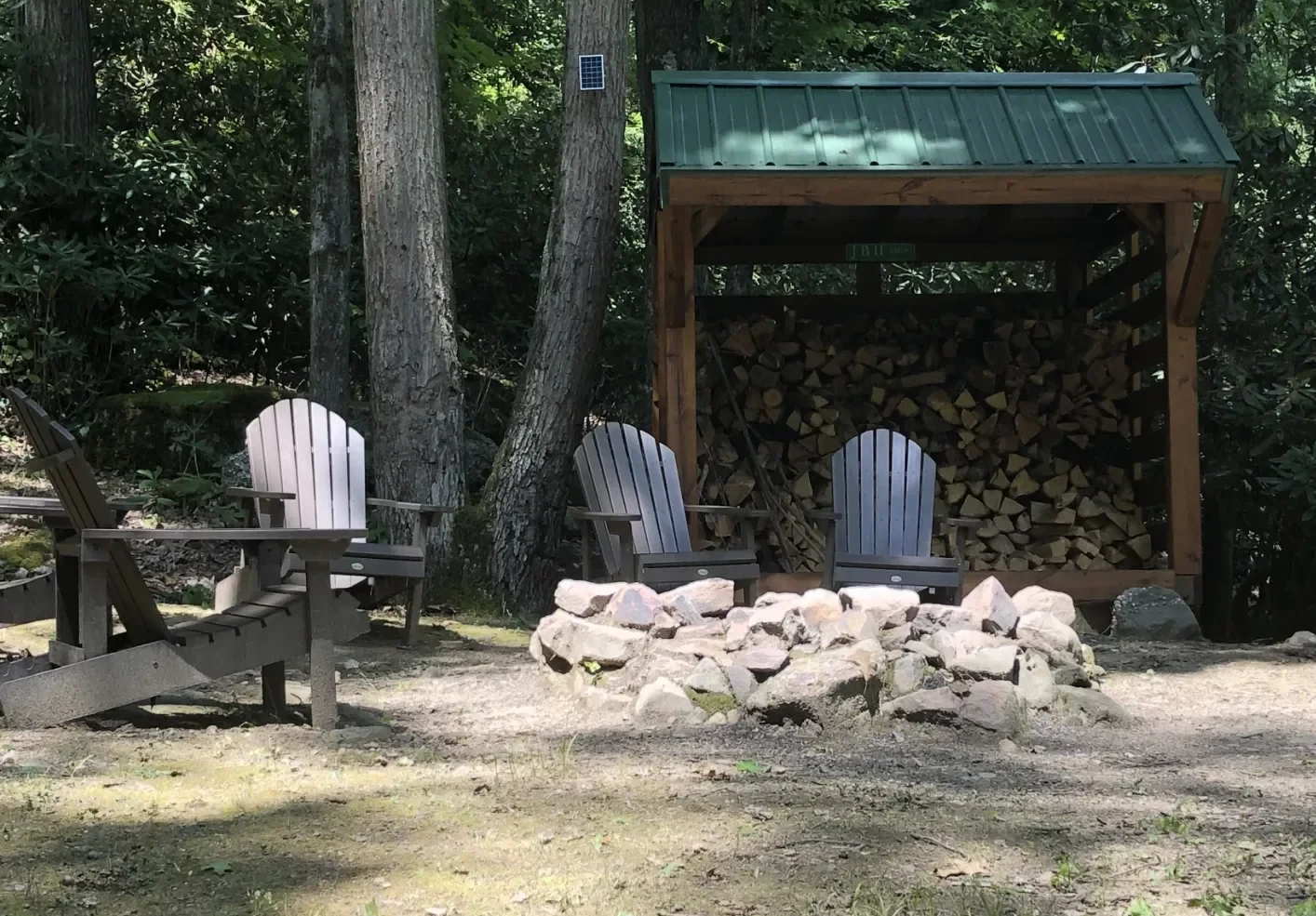 A rustic outdoor seating area in a wooded setting with three Adirondack chairs, a fire pit made of stones, and a woodpile stored under a small wooden shelter with a green metal roof. Several large trees surround the area.