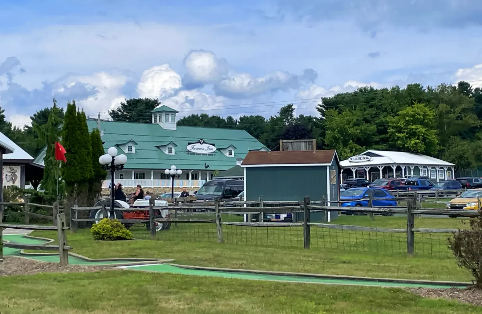 A park with a mini golf course and a parking lot in front of a building labeled 'Farmer's Inn.' There are several cars parked, and people are playing mini golf near the lamp posts. The scene is set under a partly cloudy sky with trees in the background.
