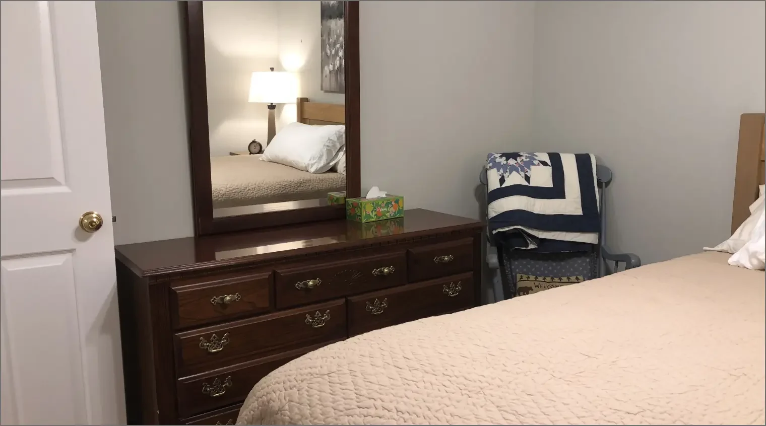 A bedroom with a dresser, mirror, bed, and a folding chair with a quilt draped over it.