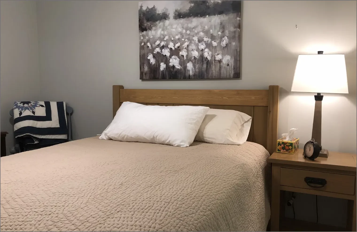 A neatly made bed with a quilted beige bedspread, white pillows, and a wooden headboard, in a bedroom with a nightstand, a lamp, a tissue box, an alarm clock, and a painting of white flowers above the bed.