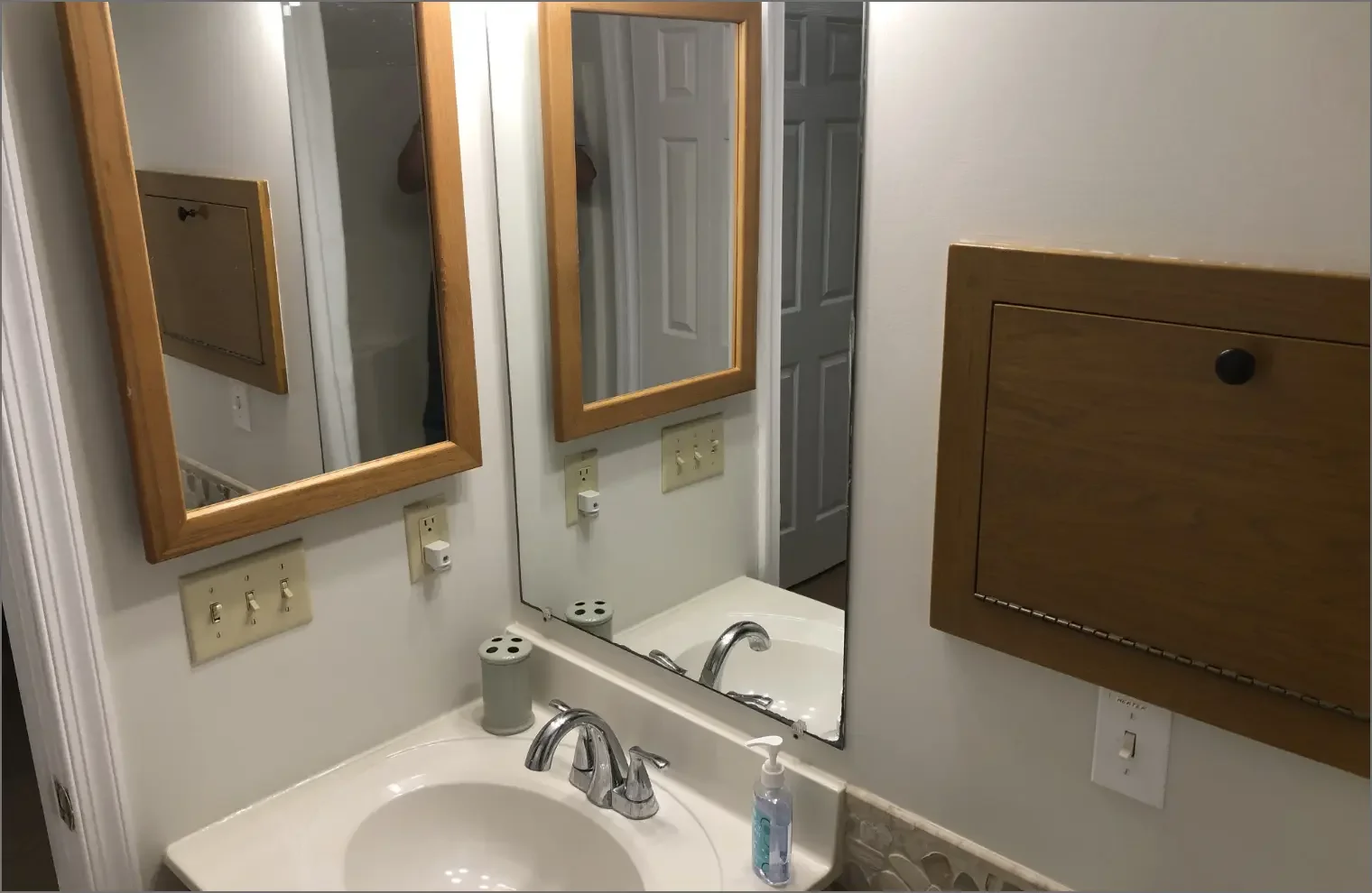 Bathroom sink with a mirror, soap dispenser, electrical outlets, a wooden medicine cabinet, and a door in the background.
