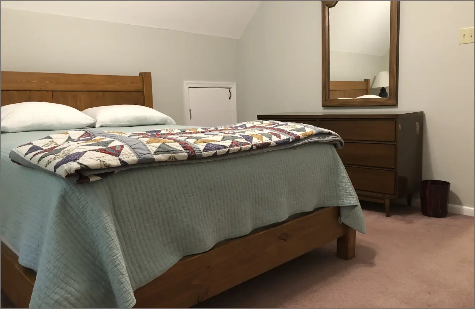 A neatly made bed with a wooden headboard, two white pillows, a light green bedspread, and a quilted blanket with a pattern, situated in a cozy bedroom with a mirror on a wooden dresser, a small door in the wall, and a waste bin on the beige carpet.