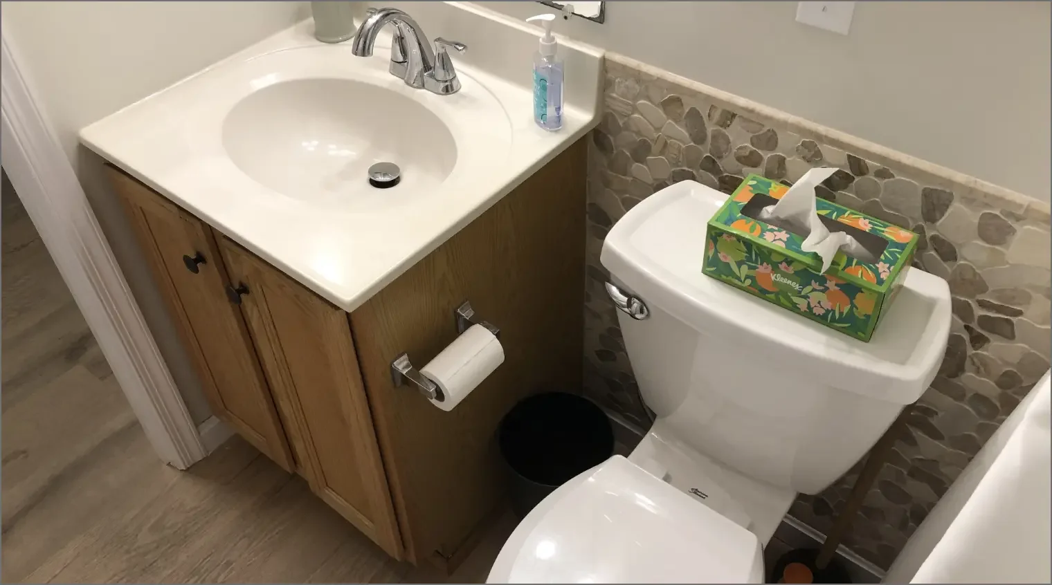 A small bathroom with a white sink, wooden cabinet, and a toilet with a box of tissues on top. There is a soap dispenser on the sink, a toilet paper roll on a holder, and a small black wastebasket on the floor.