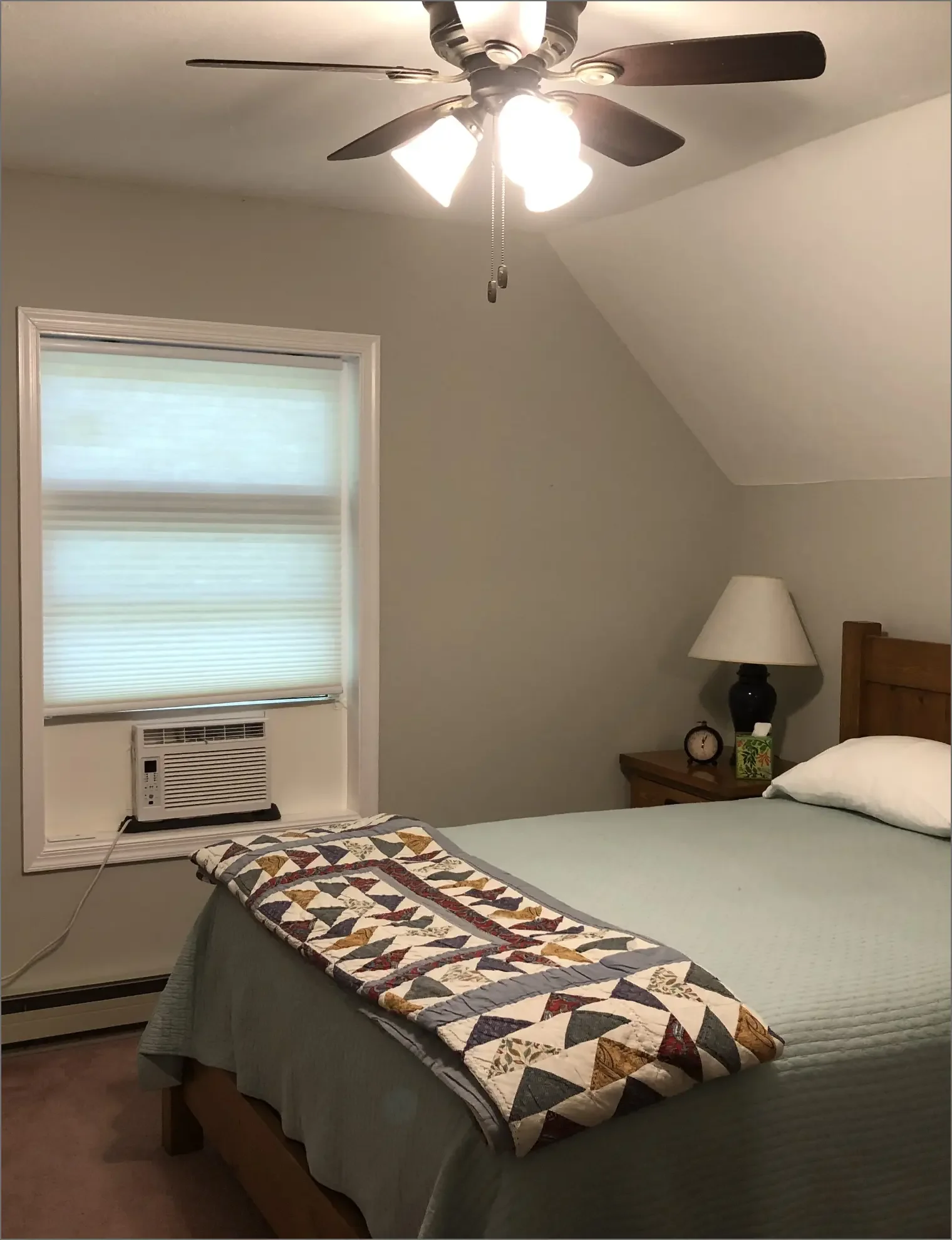 A bedroom with a bed, a wooden nightstand with a lamp, a small clock, and a tissue box. There is a window with a white blind and an air conditioning unit. The ceiling has a fan with lights.