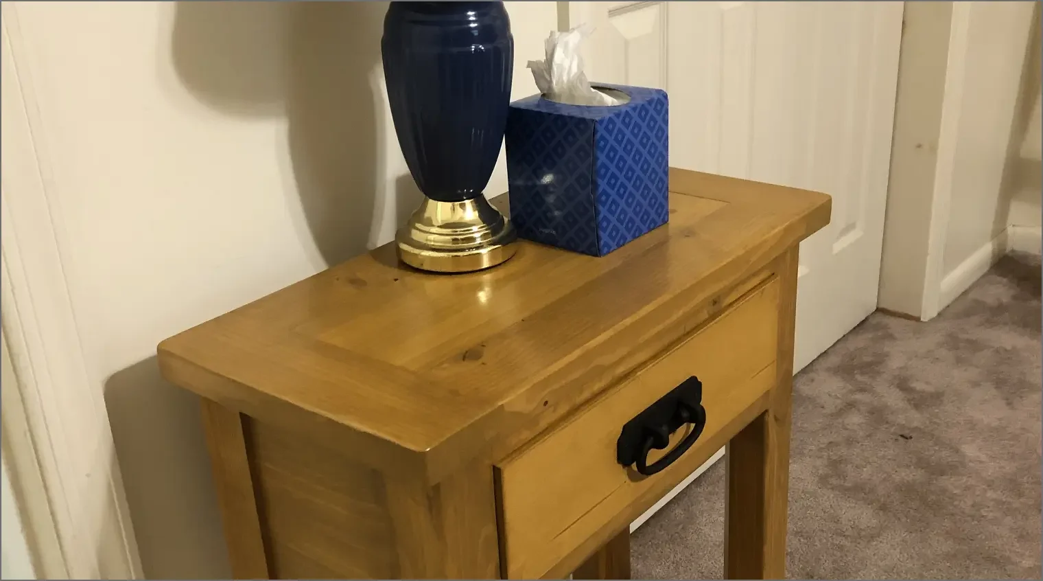 A wooden table with a blue vase with gold base, a box of tissues, and a black handle on the drawer in a room with beige carpet and white wall panels.