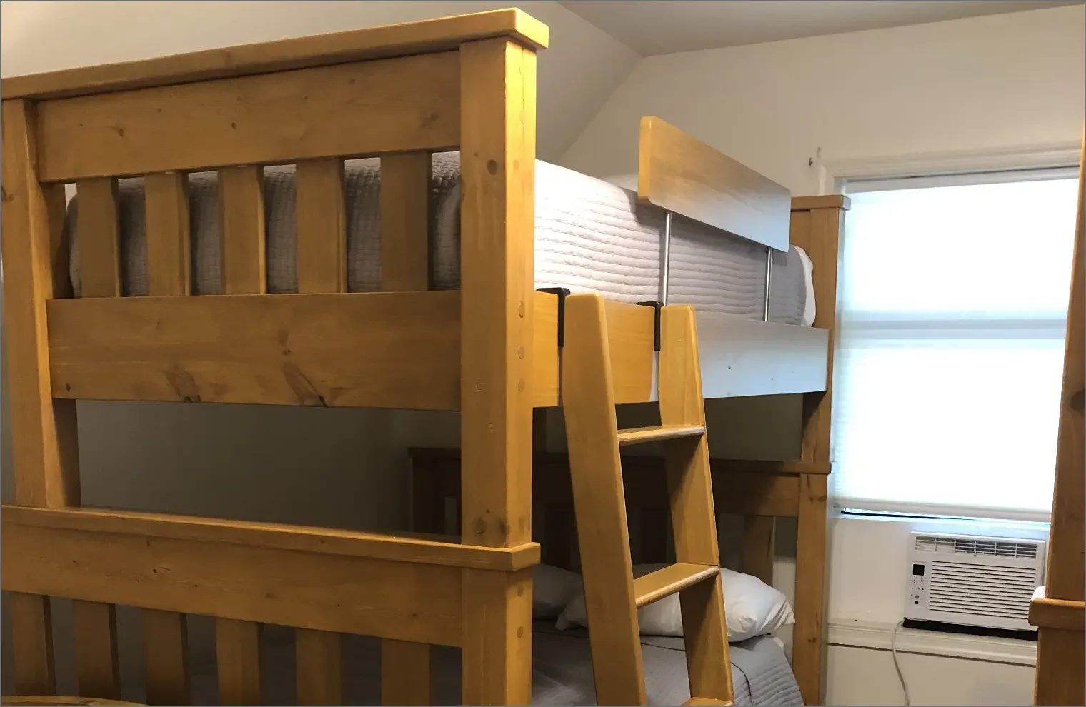 Wooden bunk beds with a small ladder, window with blinds, and an air conditioning unit in a room.