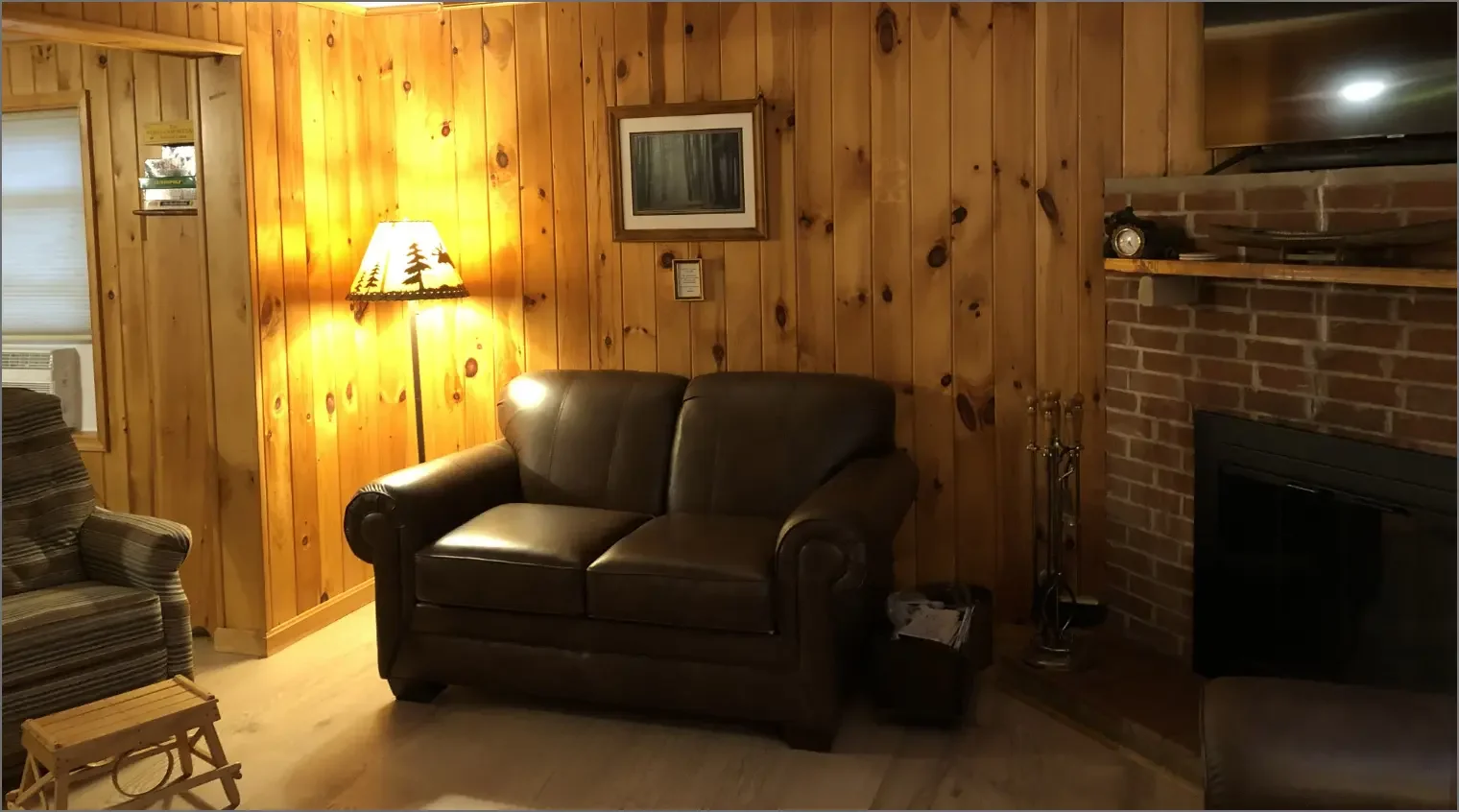 A cozy living room with wood-paneled walls, a lit floor lamp with a pine tree design, a framed picture, and a brick fireplace with tools nearby. There are dark leather and striped fabric sofas, and a small wooden bench in the foreground.