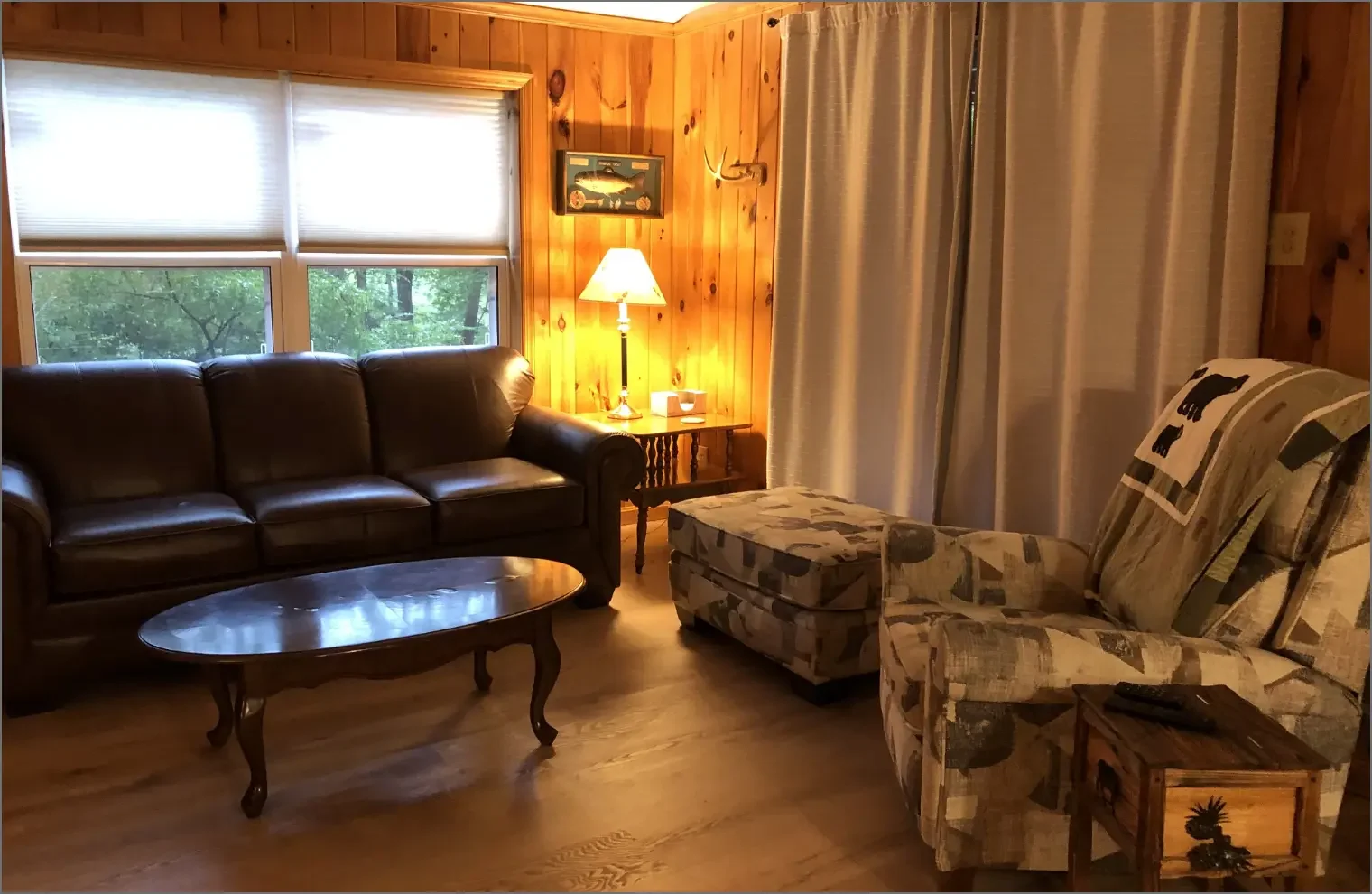 Living room with wooden walls, featuring a brown leather sofa, stained glass window, a patterned armchair with matching ottoman, side table with a lamp, and beige curtains.