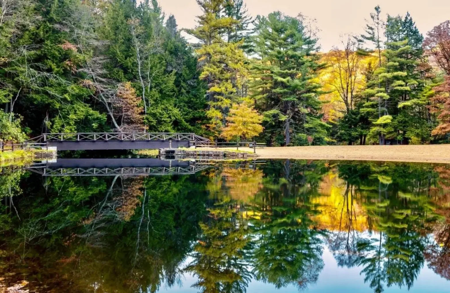 A small bridge over a calm lake surrounded by trees with fall foliage, reflecting in the water.