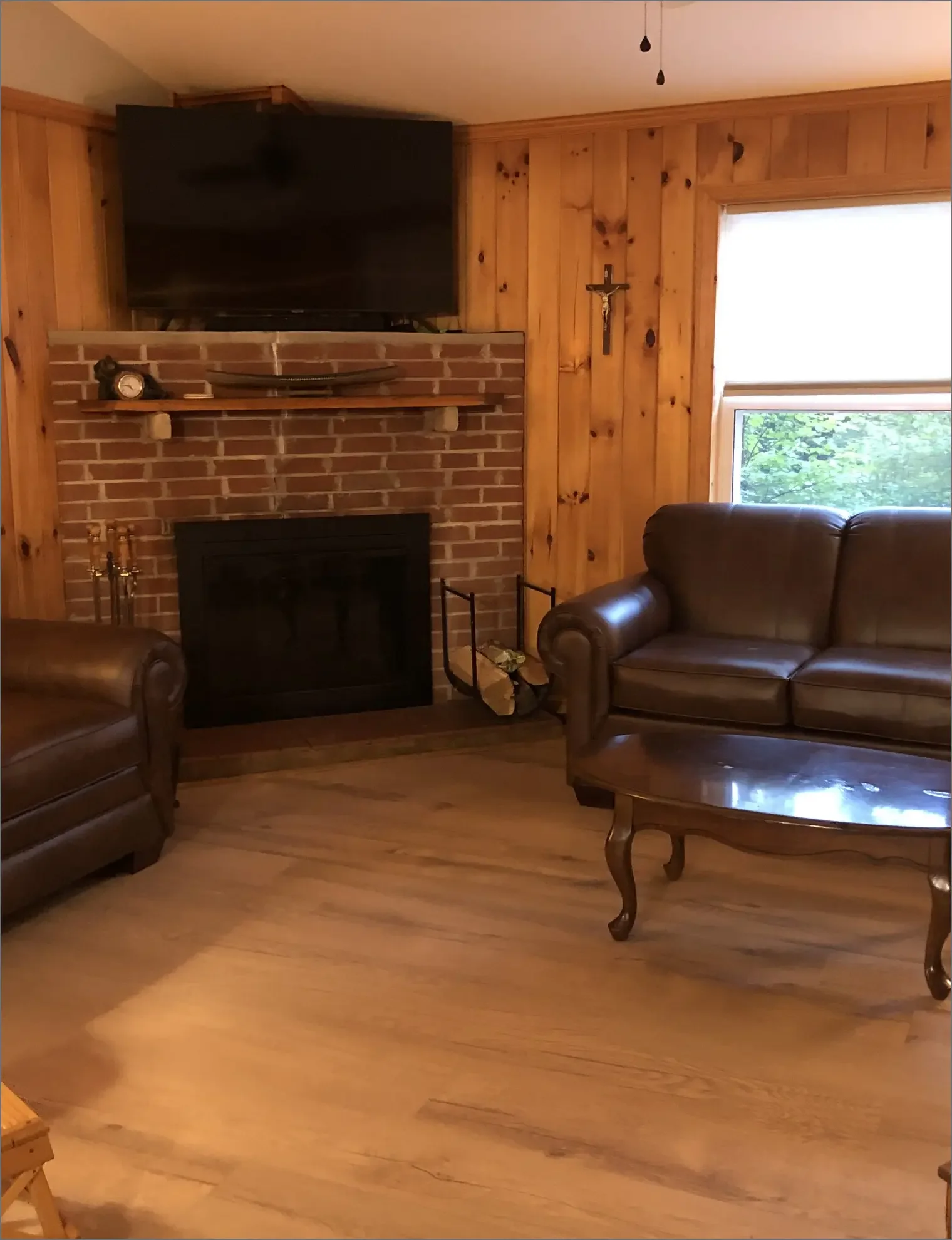 Living room with wood-paneled walls, brick fireplace, a leather sofa, a wooden coffee table, a window with a view of trees, a flat-screen TV above the fireplace, and a crucifix on the wall.