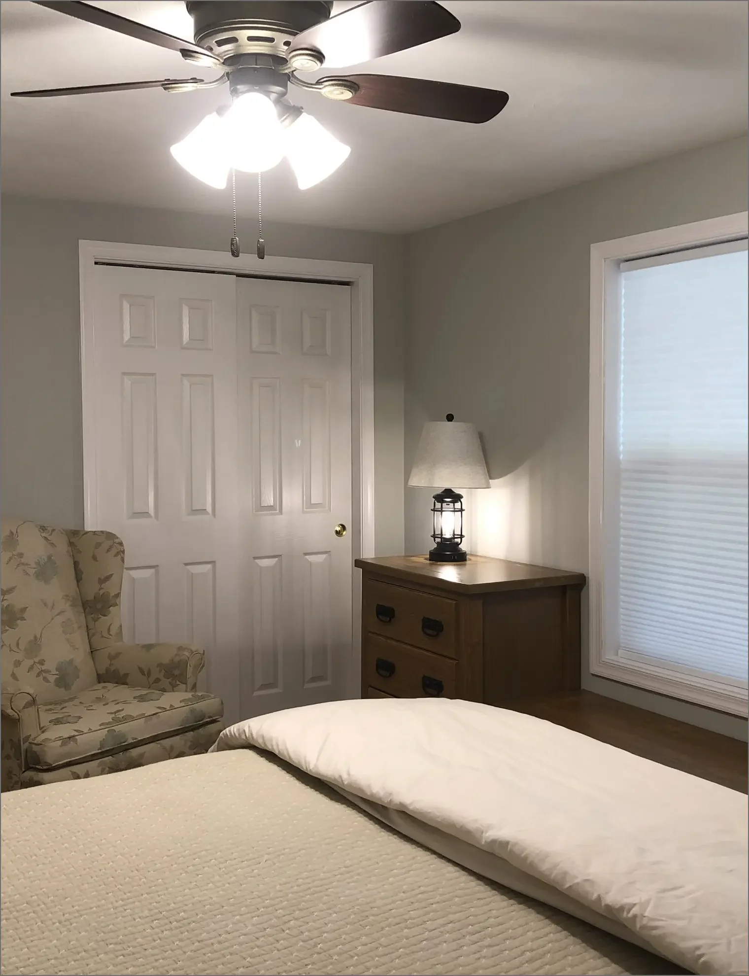 A cozy bedroom with a bed, floral armchair, wooden dresser, lamp, closet doors, and window with blinds.