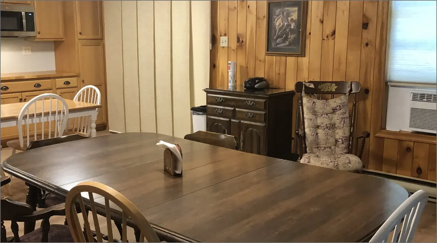 A dining room with a wooden table and mixed chairs, a wood-paneled wall, a framed picture, a chair with floral upholstery, a small dark cabinet with decorative handles, a microwave in the kitchen, and an air conditioning unit in the window.