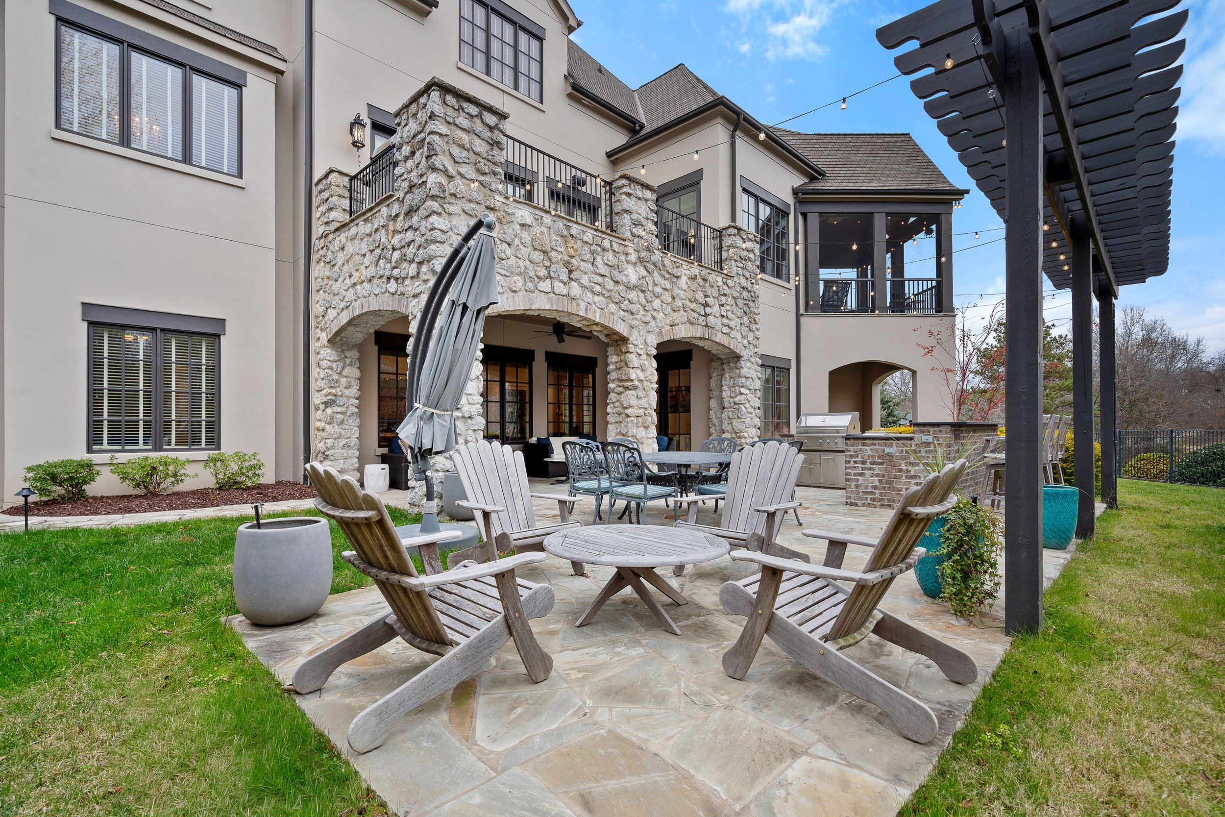 Backyard patio with wooden chairs, round table, potted plants, stone flooring, and a grill area under a black pergola, with a large house featuring stone accents and multiple windows in the background.