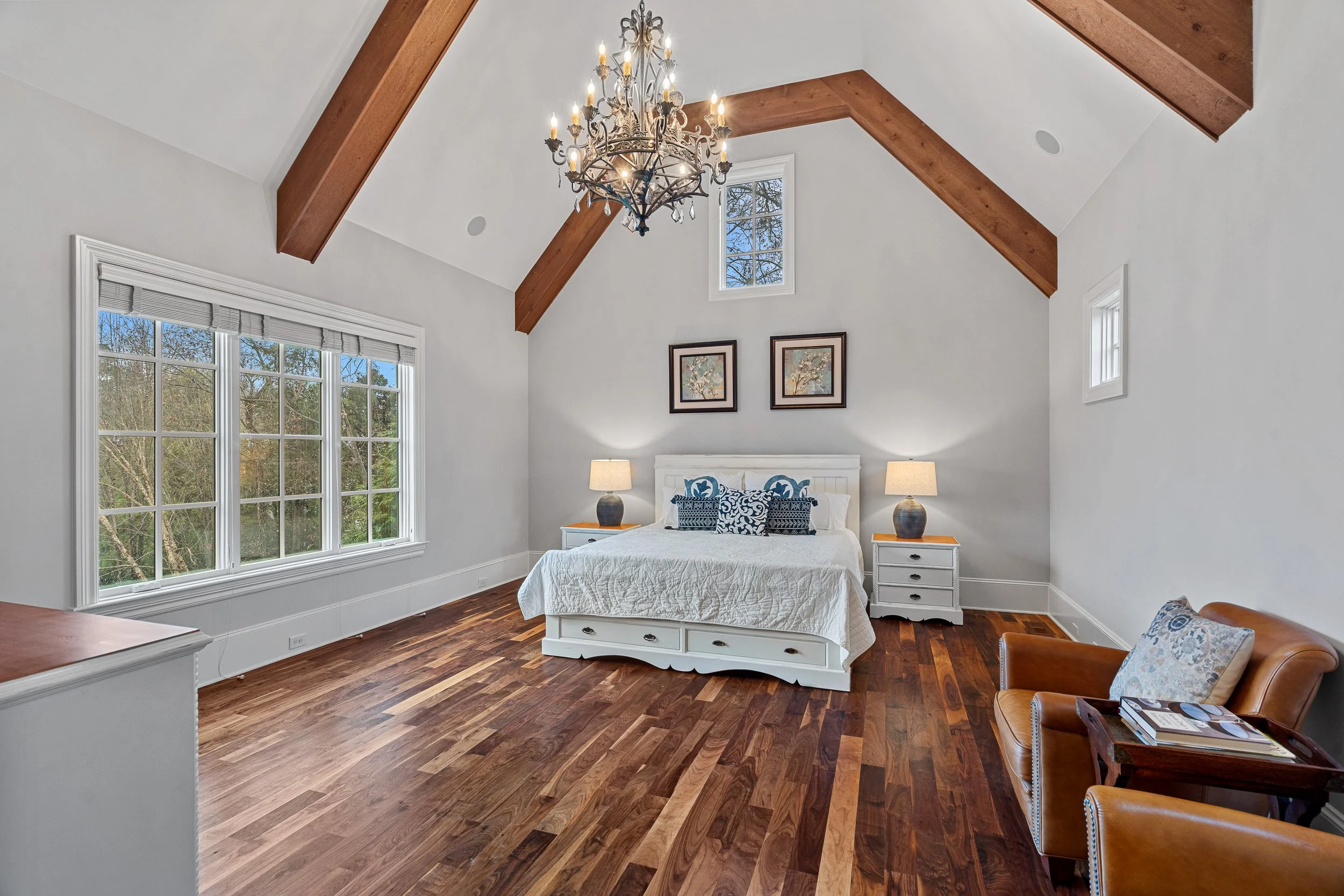 Bedroom with white bed, two nightstands with lamps, framed artwork, large window with blinds, wood ceiling beams, chandelier, brown leather chair, wood side table, hardwood floor, and painted white walls.