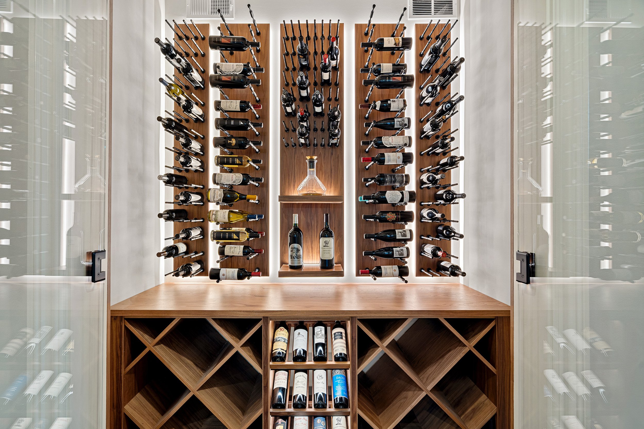 A modern wine cellar with wooden shelves holding various bottles of wine, a glass door, and a wooden counter with additional wine bottles.