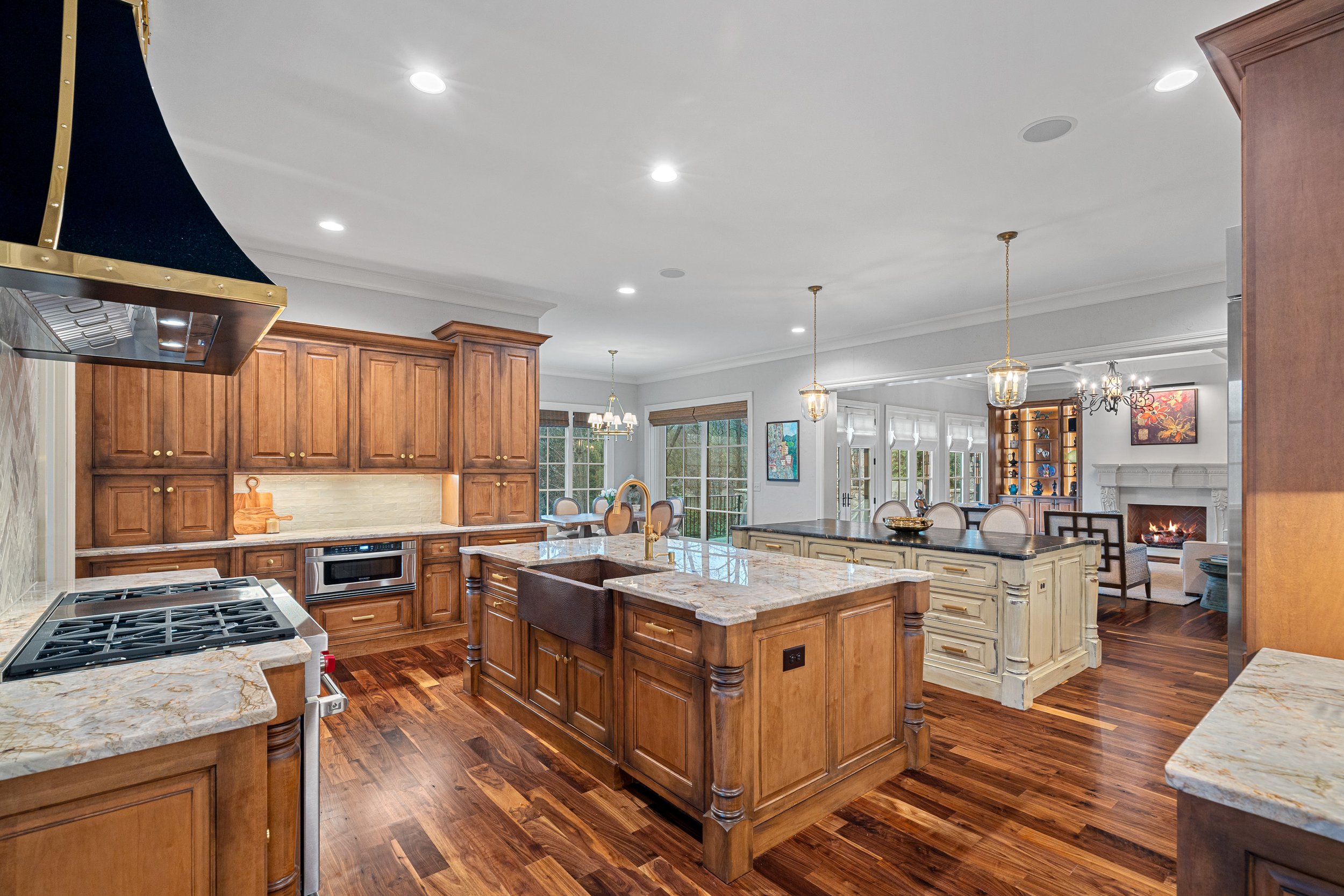 Open-concept kitchen with wooden cabinets, marble countertops, and hardwood floors, adjacent to a living area with a fireplace and large windows.