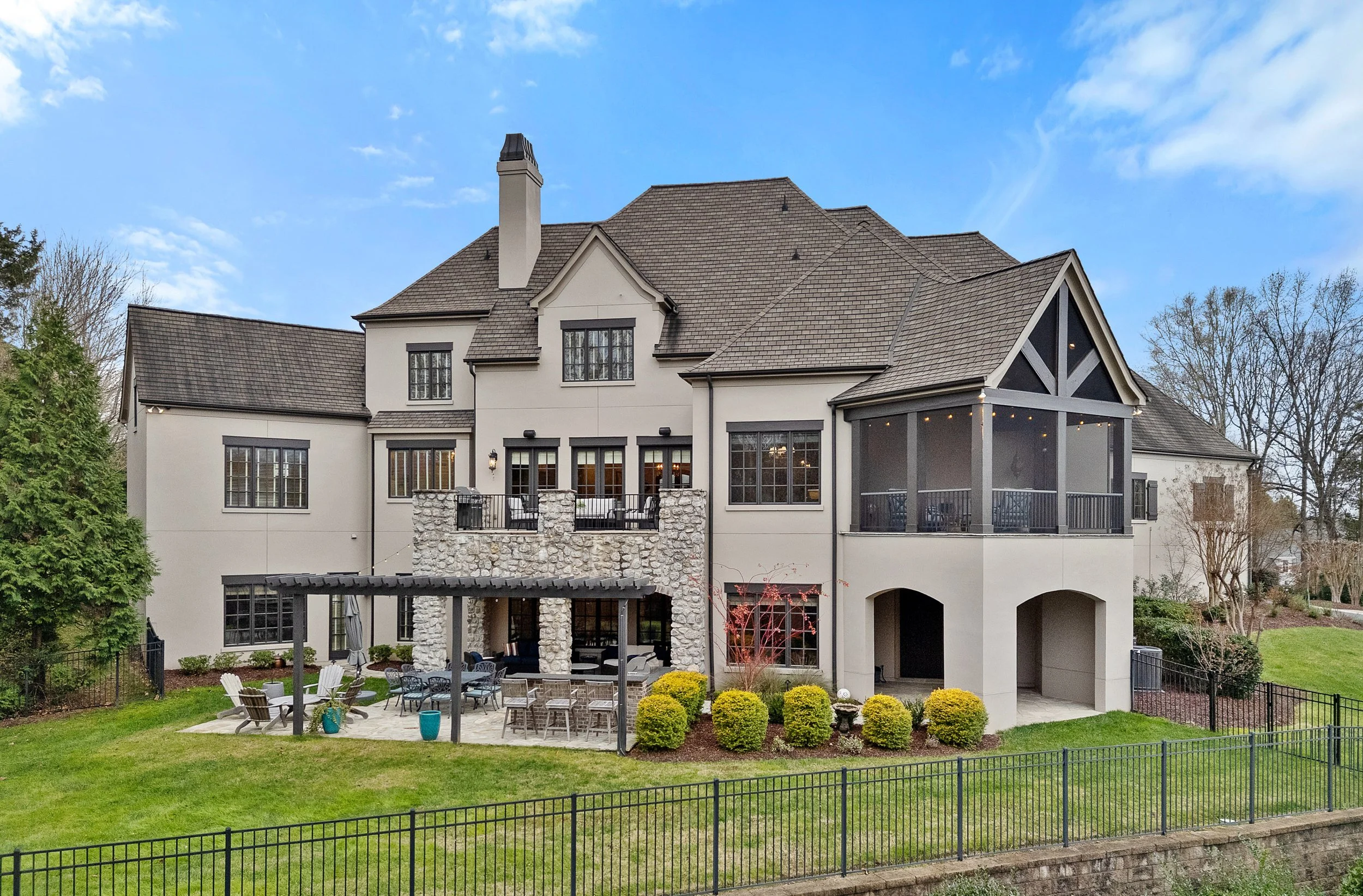 Large modern house with multiple levels, gray roof, and beige exterior. It features a screened porch with string lights, an outdoor patio with furniture, and lush green lawn enclosed by a black metal fence. Trees are visible in the background under a