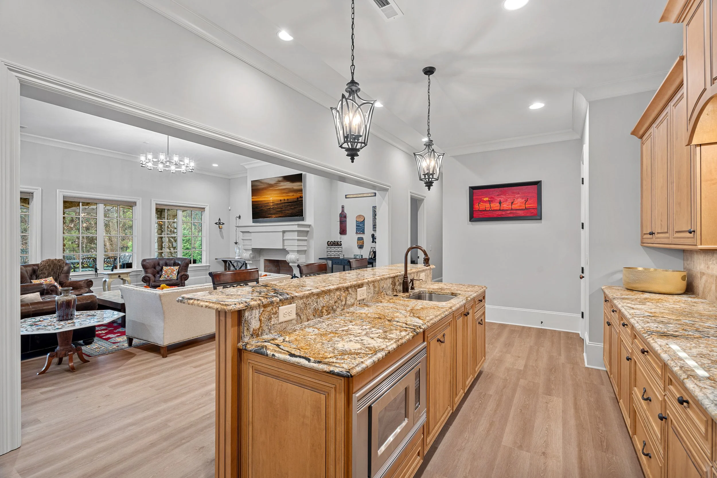 Open-concept kitchen with granite countertops and wooden cabinets, overlooking a living room with leather chairs, a fireplace, and a television.